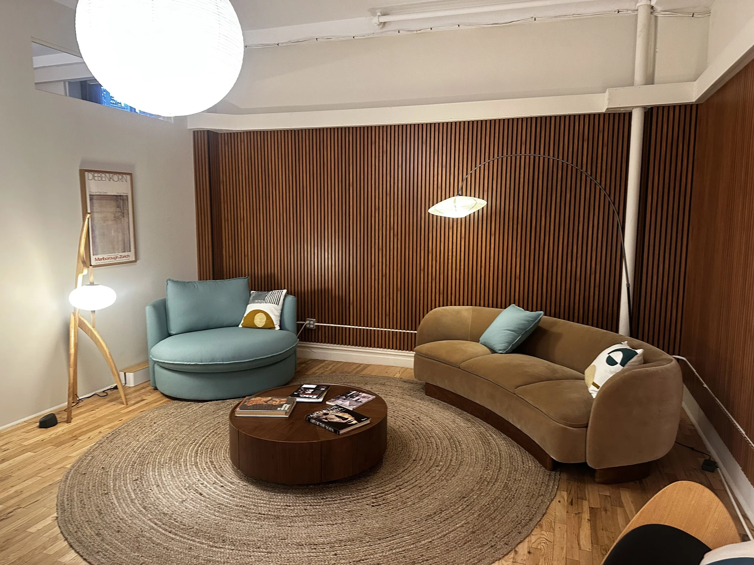 A cozy living room with a curved beige sofa, a light blue armchair with a throw pillow, a round wooden coffee table with magazines, a textured round rug, a wooden accent wall, a floor lamp, and a blue and white table lamp. The room has wooden flooring and a light-colored wall with a framed poster.