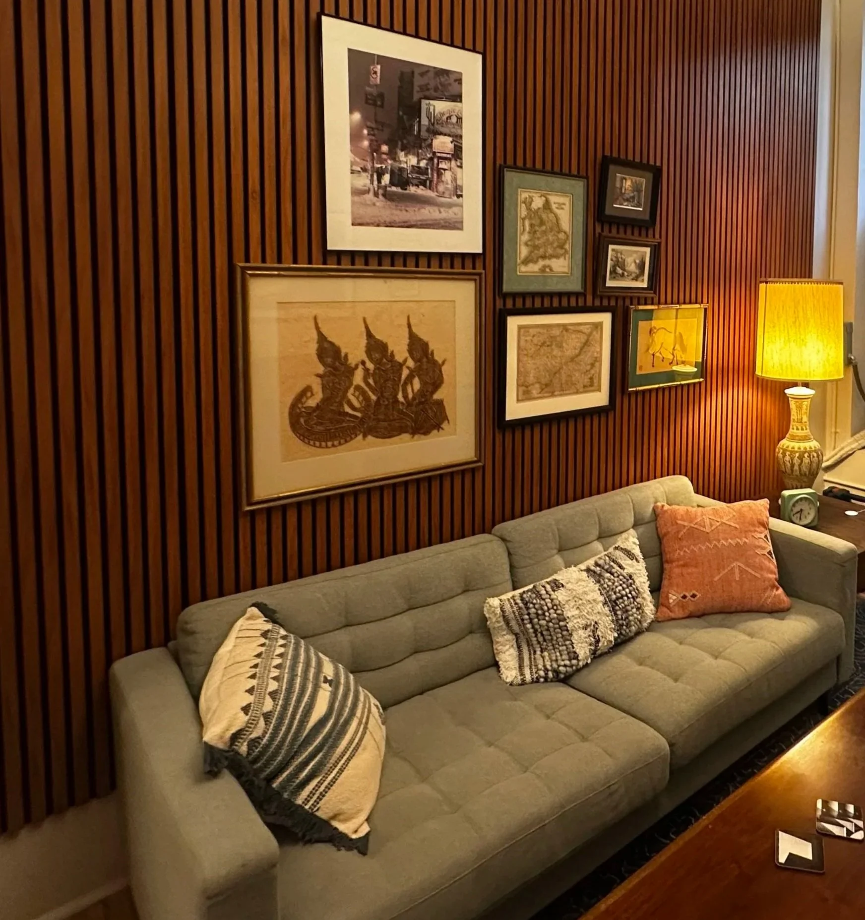 A gray tufted sofa with patterned pillows on a wooden floor beside a wooden-paneled wall with framed artwork and a yellow table lamp