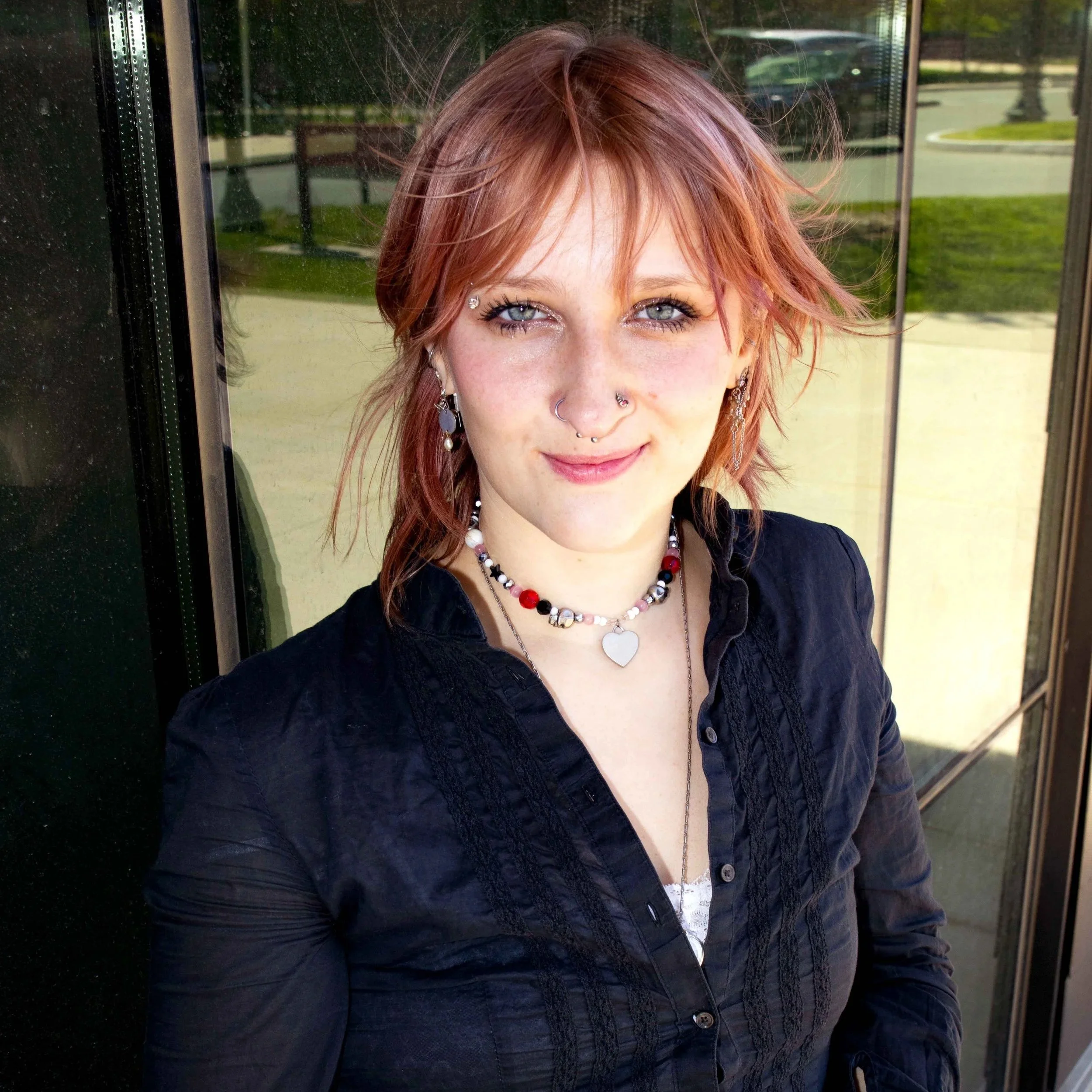 Close-up of a woman with red hair, blue eyes, and multiple facial piercings, wearing jewelry, standing outside near a reflective glass window.
