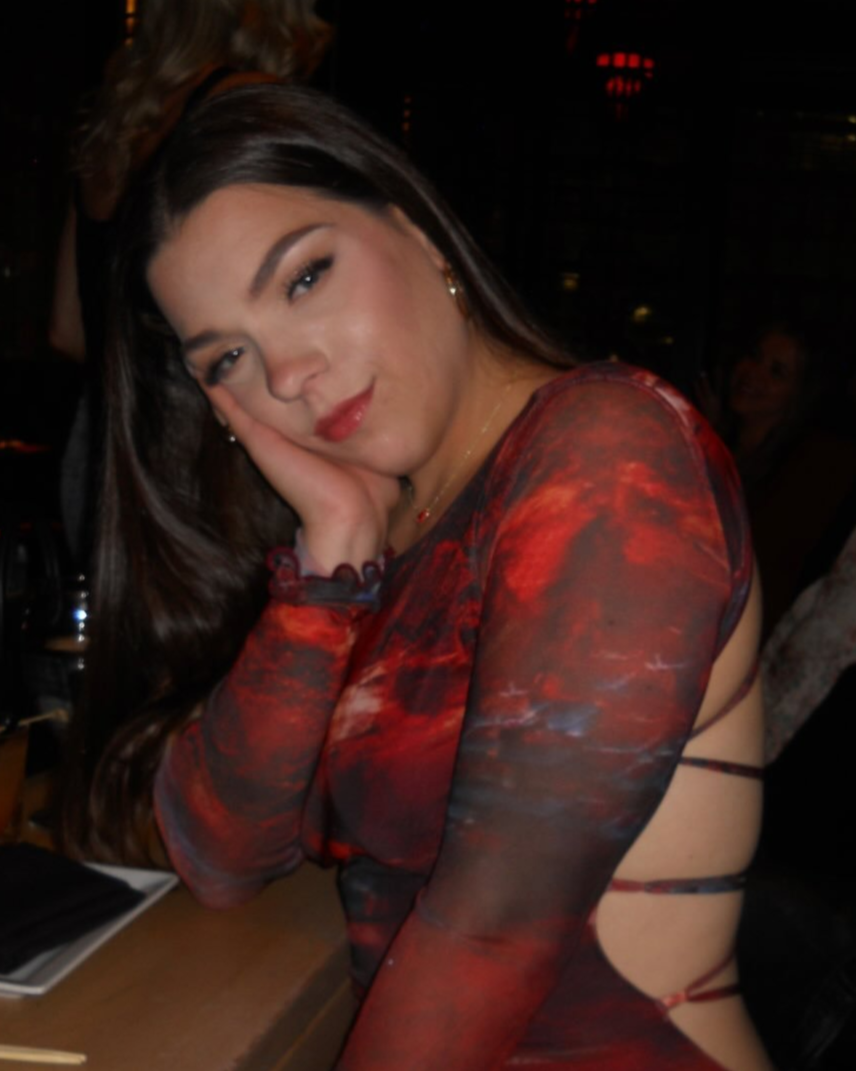 A young woman with long dark hair and light makeup, wearing a red and black patterned dress, resting her face on her hand at a dimly lit restaurant or bar.