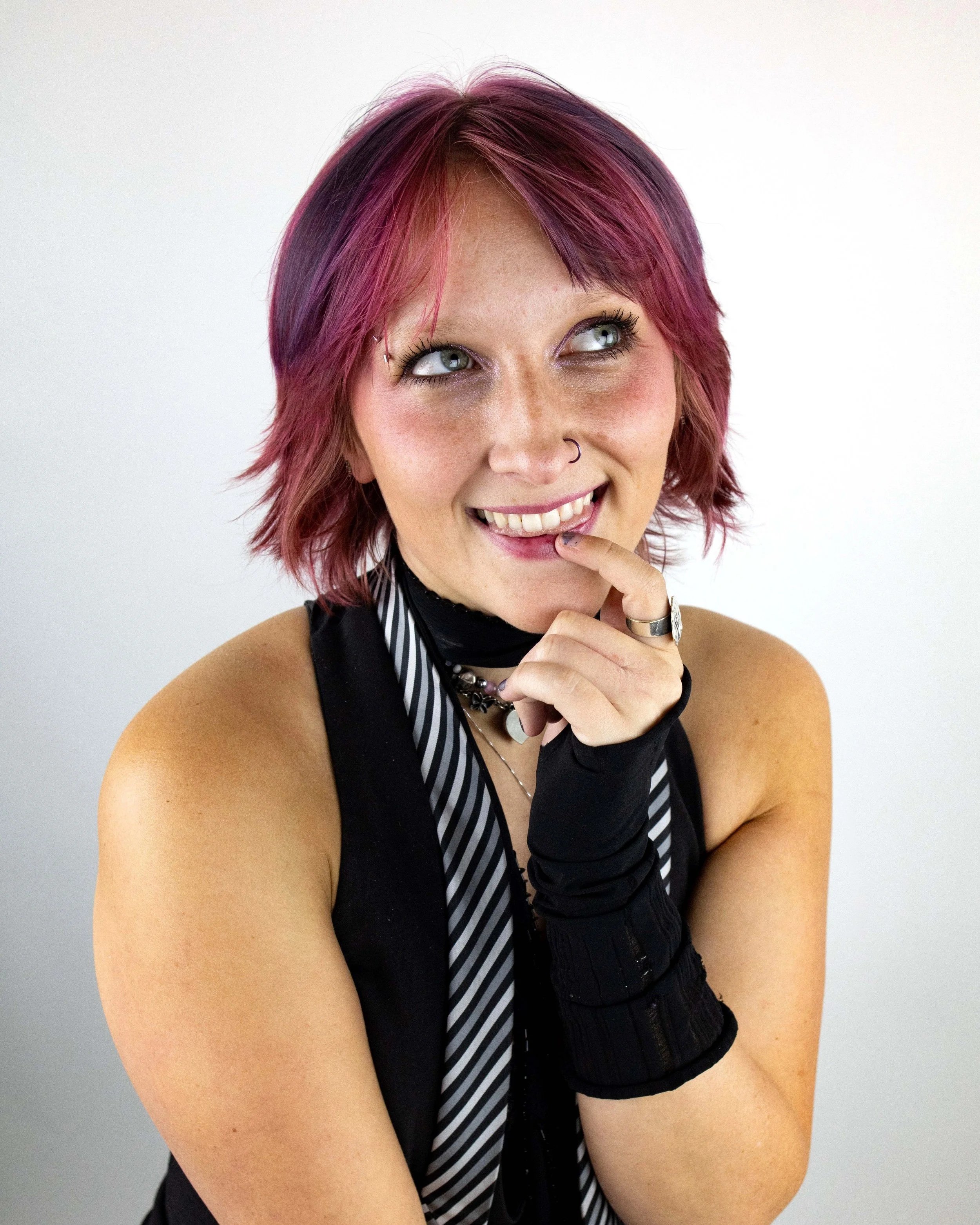 Portrait of a woman with short, layered, pinkish-purple hair, wearing makeup, a nose ring, multiple necklaces, and black gloves, smiling with her teeth showing and touching her chin.