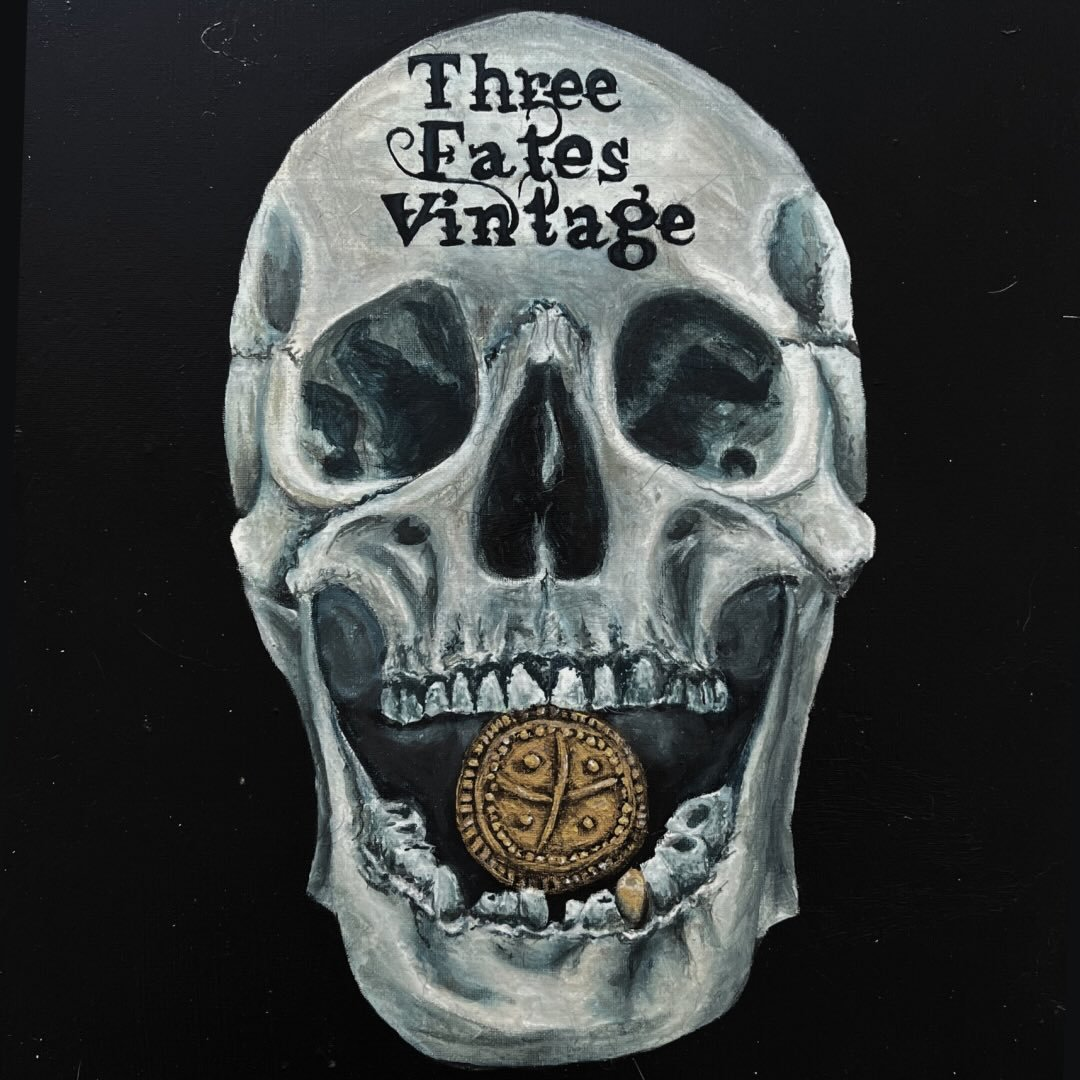 A painted skull with the words 'Three Fates Vintage' on the forehead and a gold pendant with a tree symbol in the mouth.