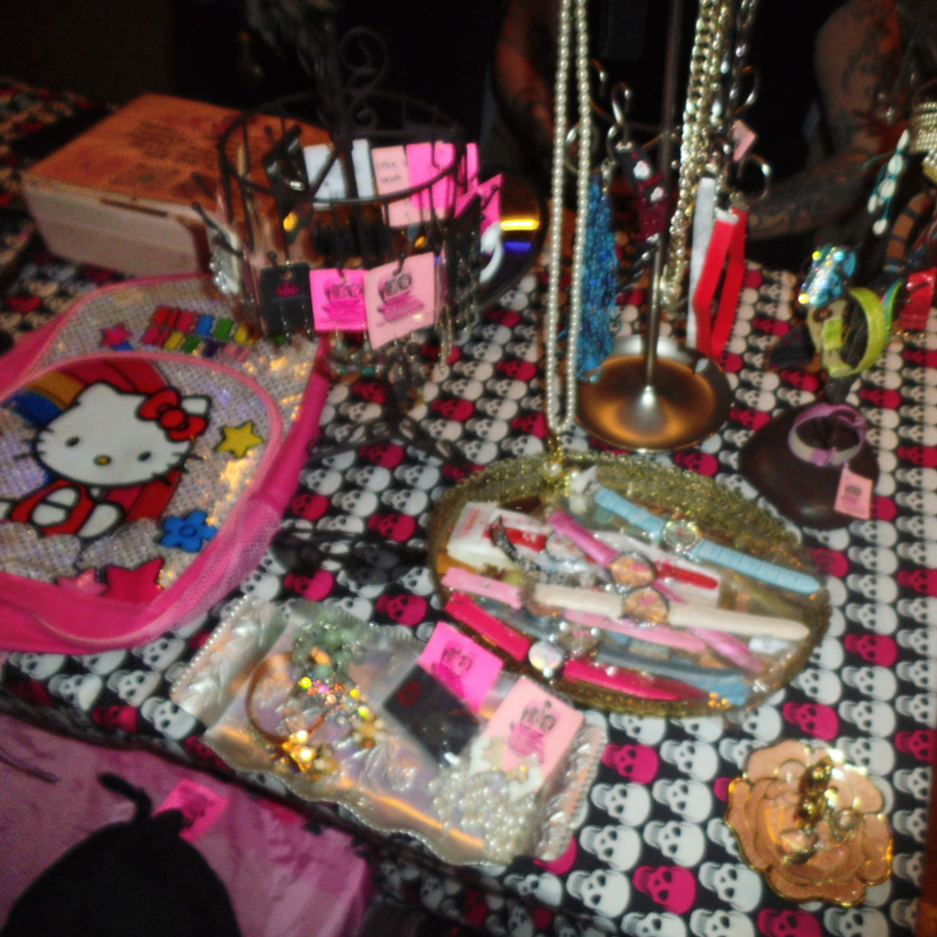 A display table featuring jewelry and accessories, including necklaces, bracelets, watches, and hair clips, adorned with pink tags and set against a black and white skull patterned tablecloth.