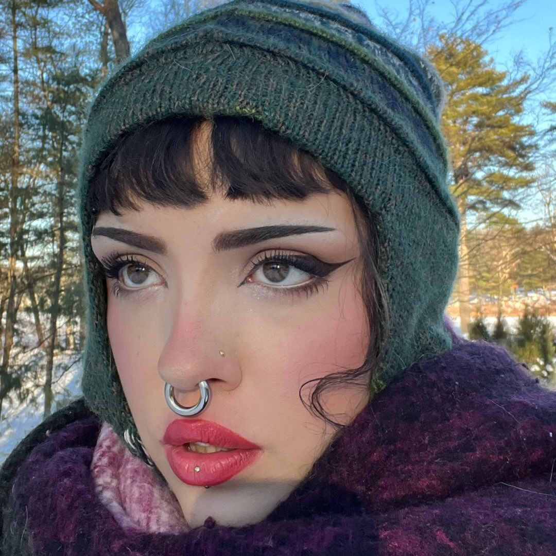 A woman with dark hair, bold makeup, and multiple facial piercings, wearing a knit hat and scarf, outdoors with trees and a winter landscape in the background.