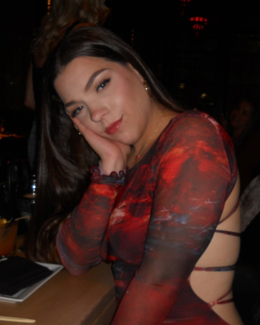 A young woman with long dark hair and makeup, resting her face on her hand and smiling slightly, wearing a red and black patterned dress with cut-outs on the side, sitting indoors at a bar or restaurant.