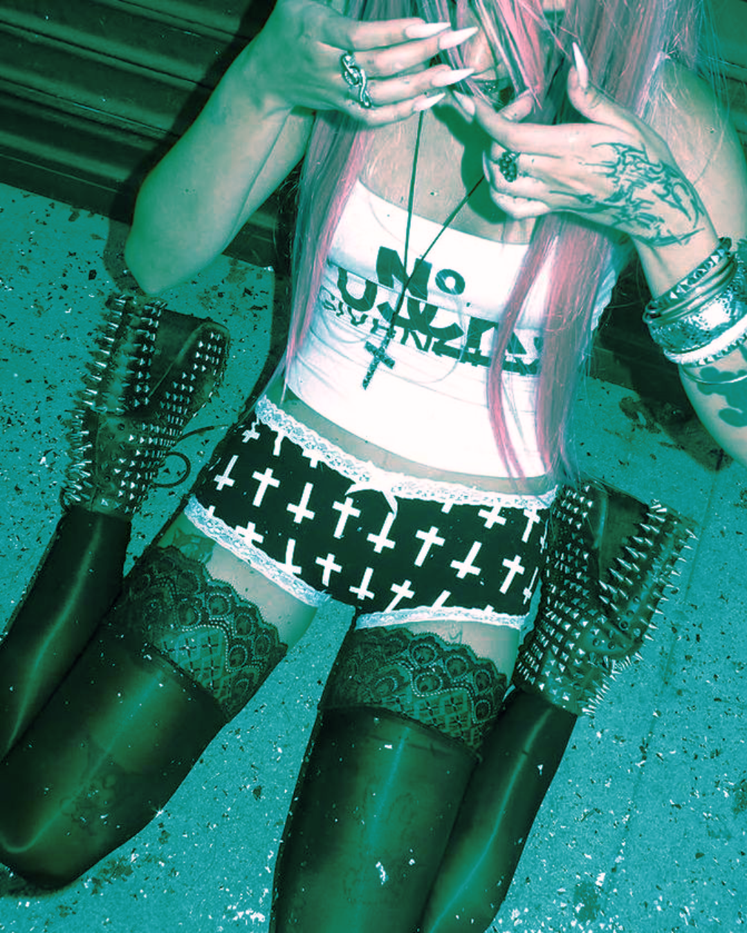 A woman with pink hair and tattoos, wearing a white tank top with black lettering, patterned shorts with crosses, black lace stockings, and thigh-high black boots with spikes, sitting on a gravel surface with her knees bent and hands near her face.