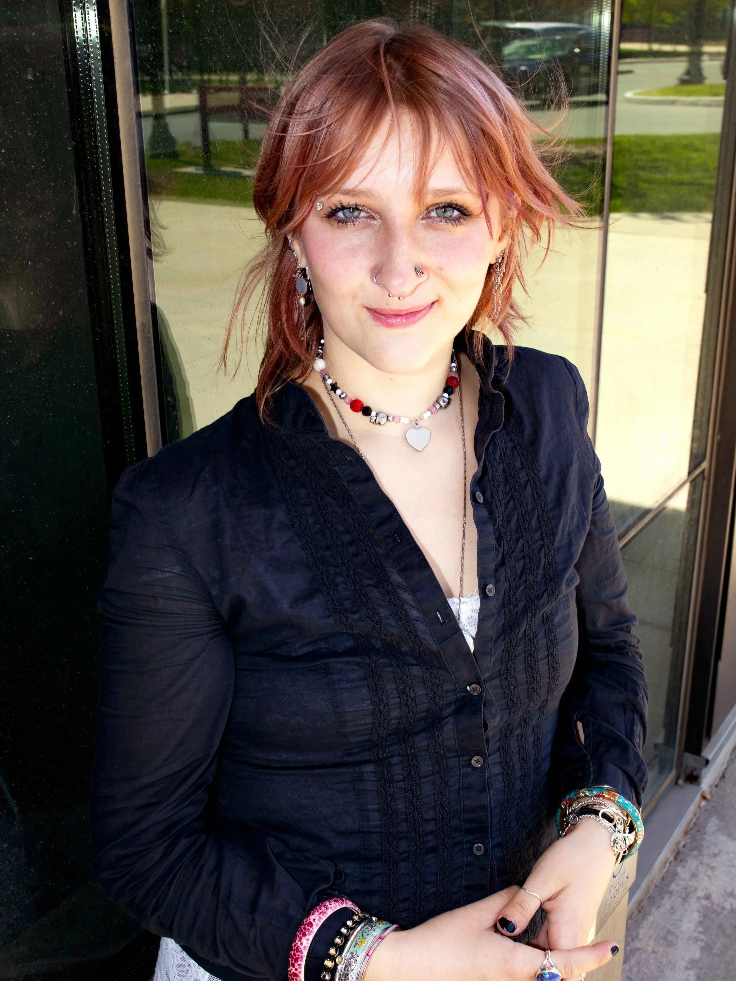 A young woman with reddish hair, blue eyes, and multiple facial piercings smiling outdoors. She is wearing a black shirt, colorful jewelry including bracelets and necklaces, and multiple earrings. Her hands are clasped in front of her, showing additional jewelry. The background shows a glass window and a park or street scene.