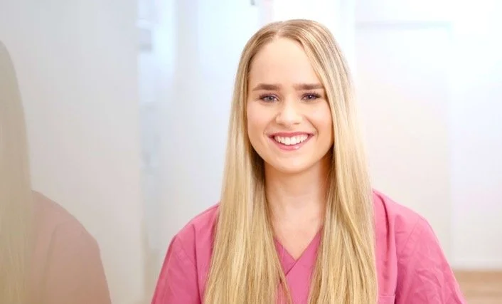 Eine junge Frau mit langen blonden Haaren, die ein pinkes Pflegeoutfit trägt, lächelt freundlich in die Kamera.
