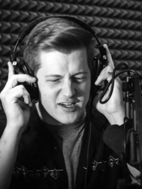 Young man singing into a microphone in a recording studio with soundproofing panels, wearing headphones and expressing emotion.