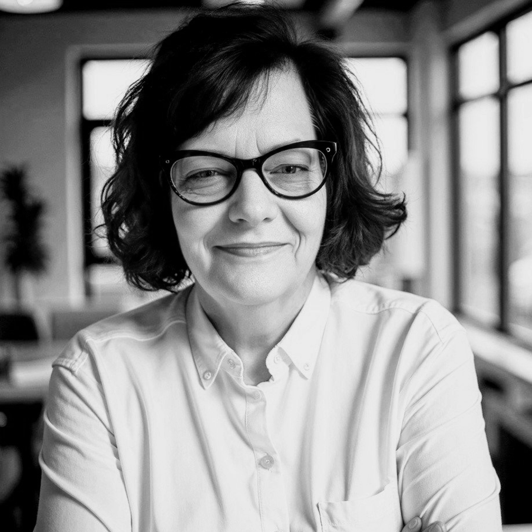 Black and white photo of a woman with short wavy hair and glasses, smiling, indoors with large windows and blurred background.