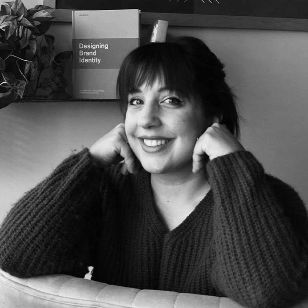 A woman with short dark hair smiling, resting her head on her hands in front of a book titled 'Designing Brand Identity'.
