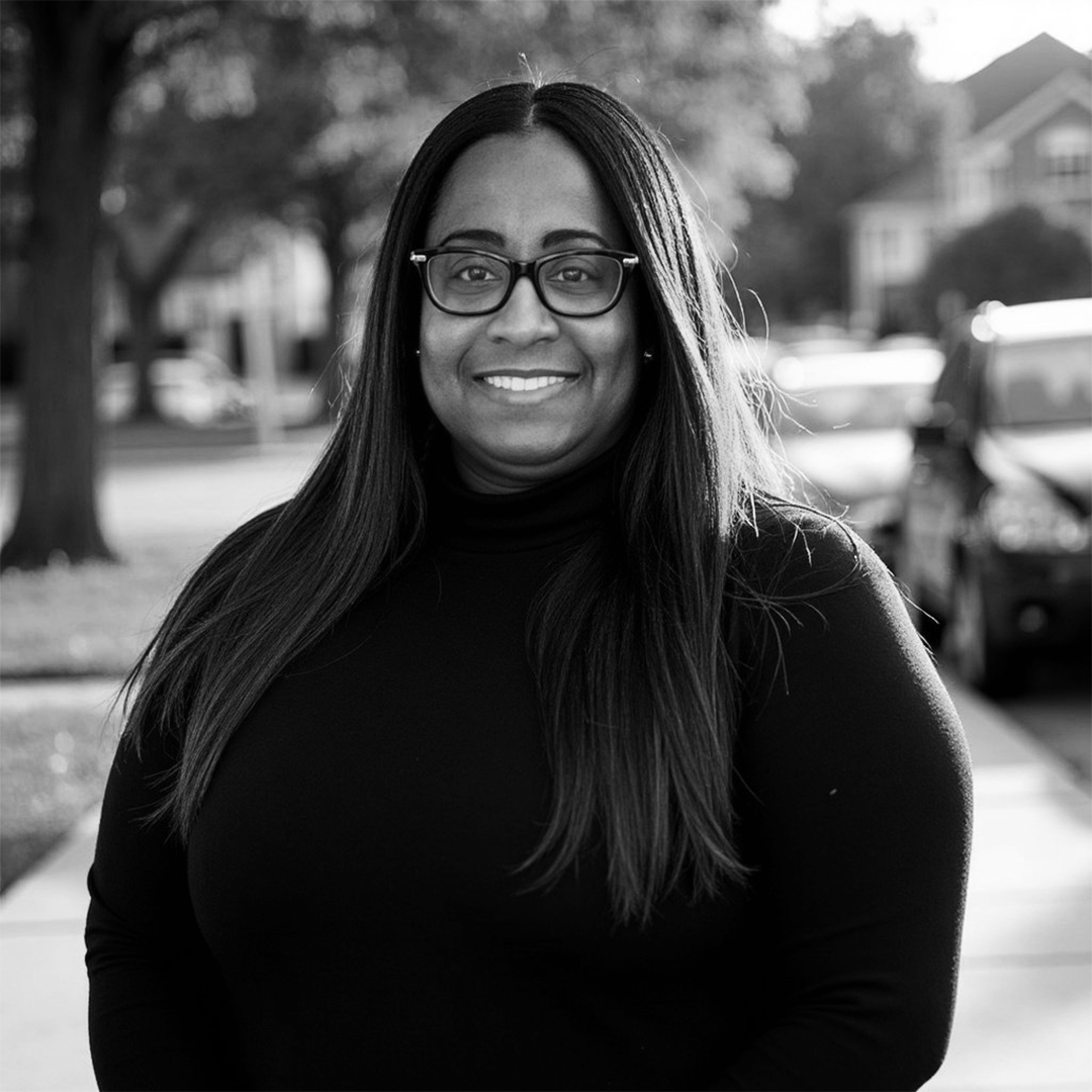 A smiling woman with long dark hair and glasses, standing outdoors on a sidewalk with cars and trees in the background, in black and white.