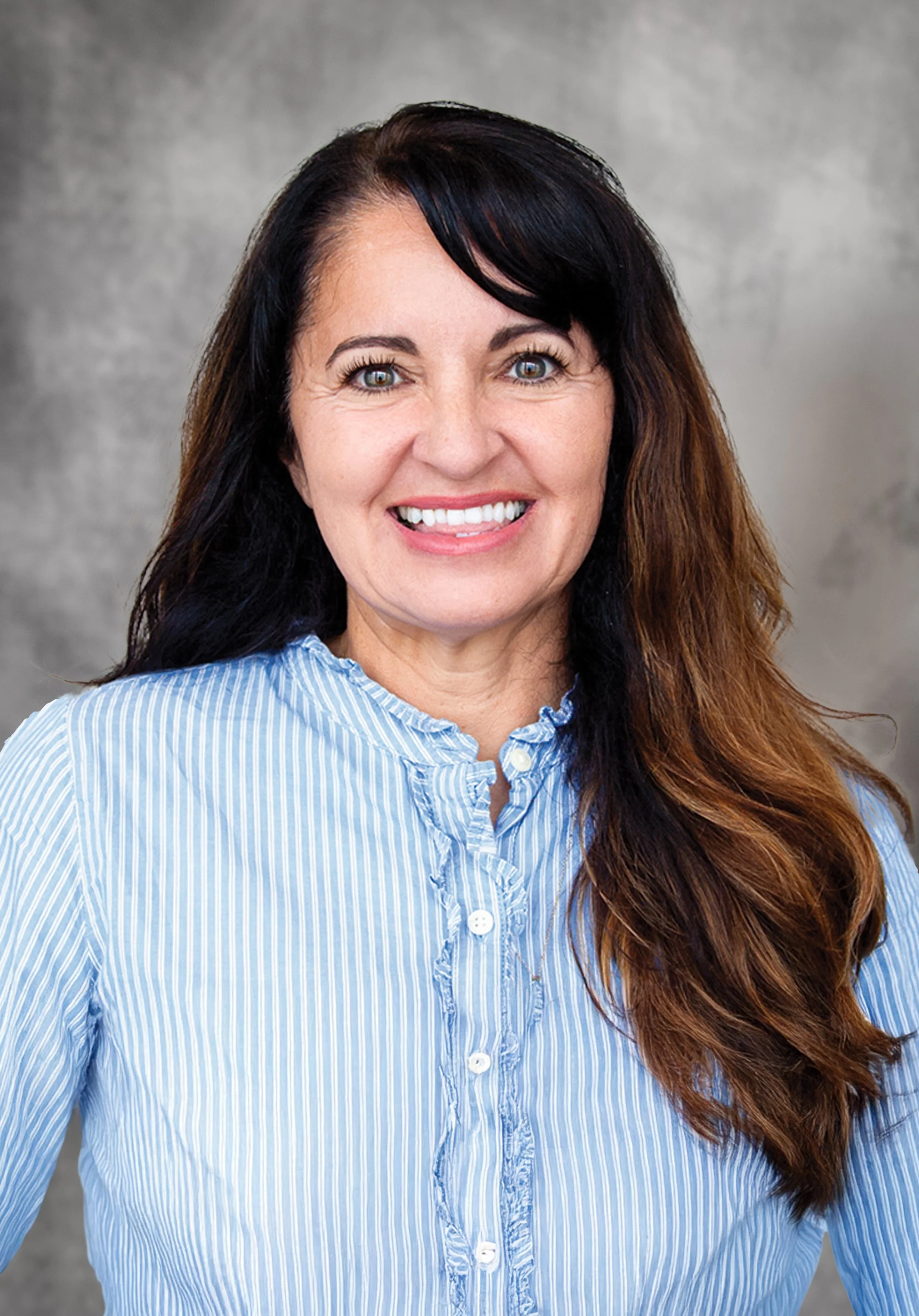 Close-up of a woman with long, dark brown hair, blue eyes, and braces, smiling and wearing a light blue and white striped button-up shirt.
