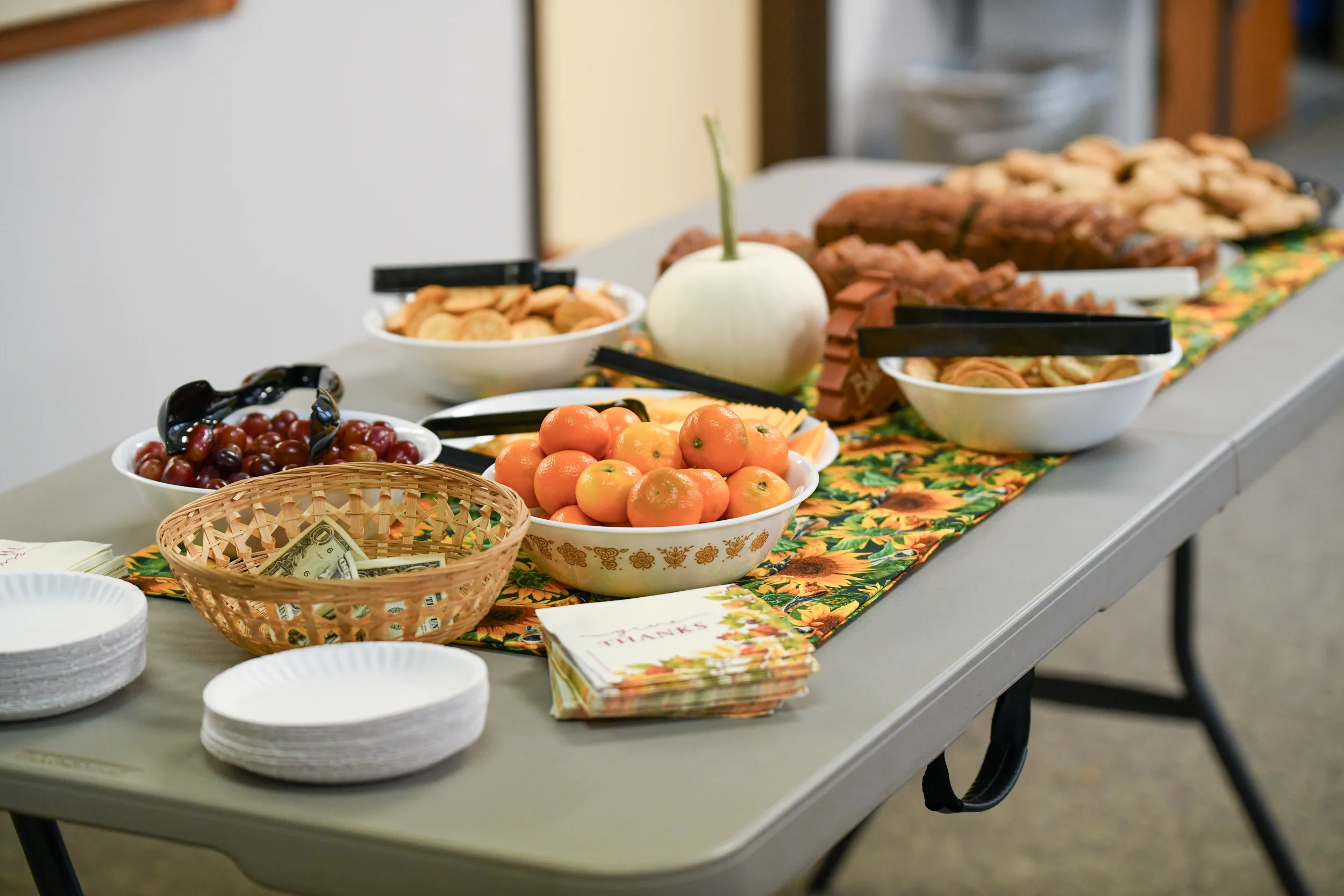 A table decorated with a festive tablecloth displays bowls of grapes, oranges, and assorted baked goods, with paper towels and a thank you card nearby.