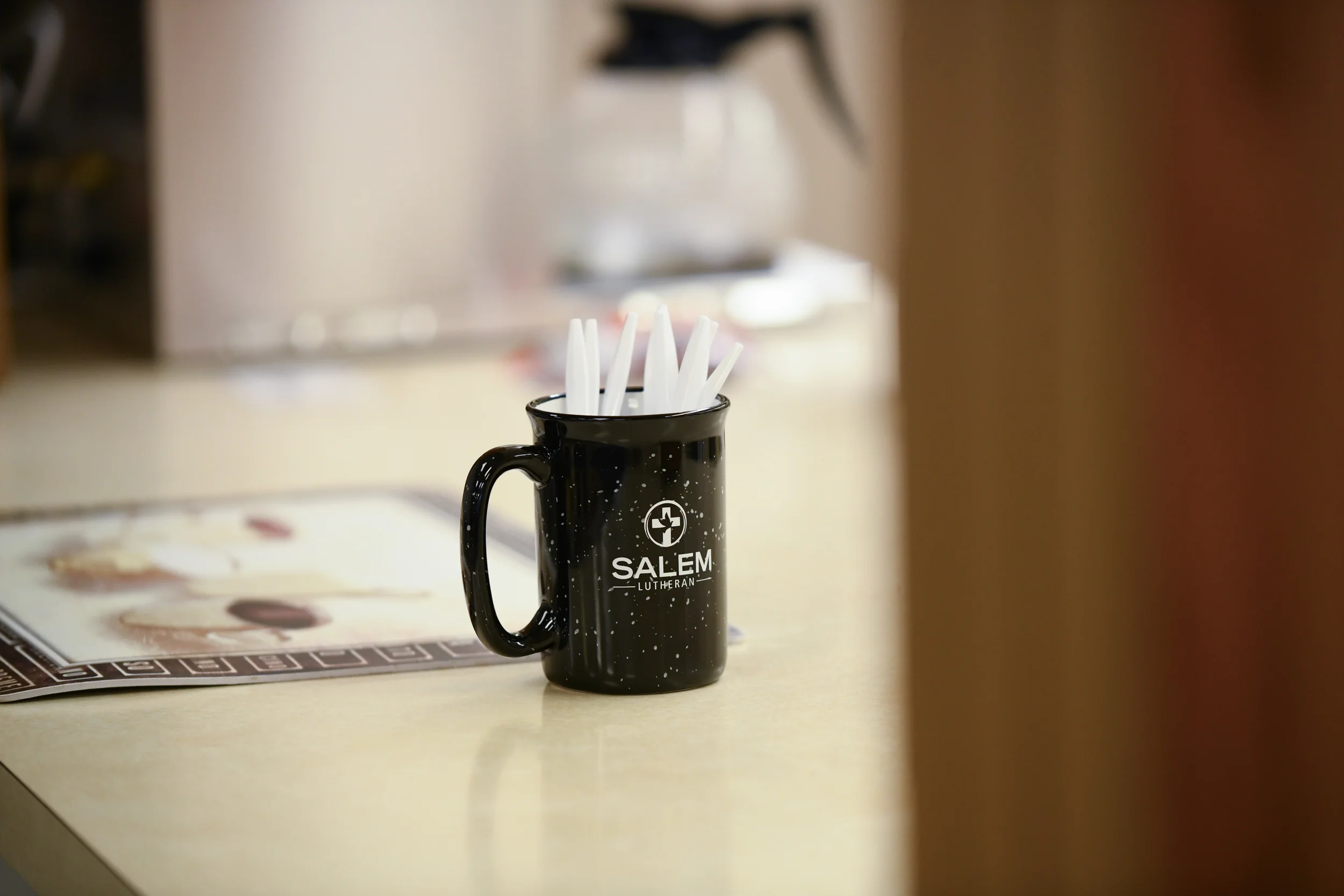 A black ceramic mug with white speckles and the words 'Salem Lutheran' printed on it, filled with white plastic stir sticks, sitting on a light-colored counter with a blurred background.