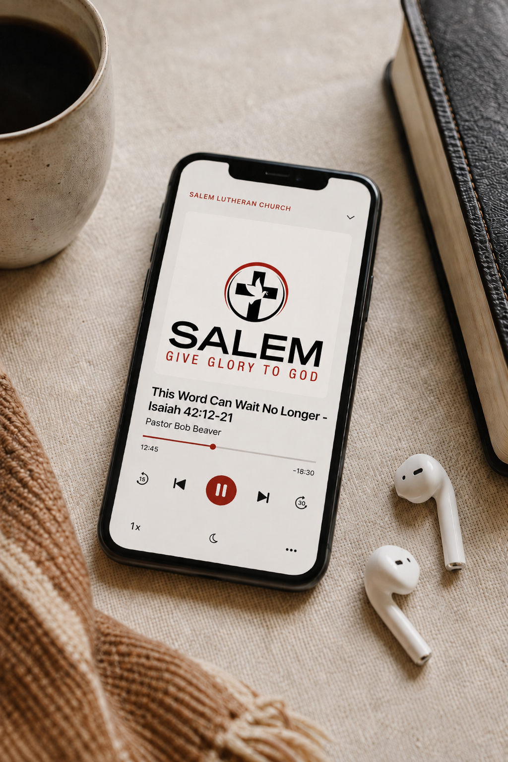 Smartphone displaying a church podcast titled 'Salem Give Glory to God' with a Bible verse from Isaiah 42:12-21, resting on a beige fabric surface along with a white coffee mug, a pair of wireless earbuds, a book, and a knitted fabric.