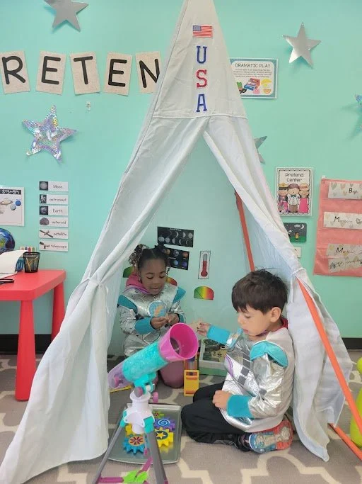 Children playing with a telescope and toy tools inside a white play tent decorated with an American flag and the word 'RETEN' on the wall, in a colorful classroom.