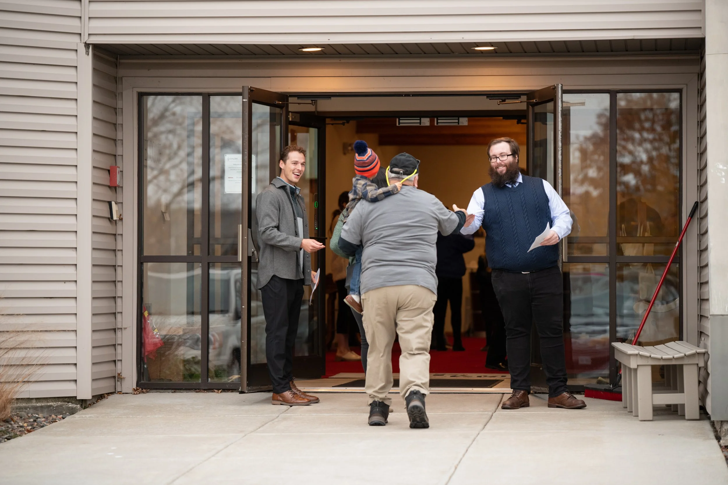 People greeting each other at the entrance of a building, including a man with glasses and a beard, a young man in a gray blazer, and a man with a child on his back wearing a winter hat. The scene appears to be friendly and informal.