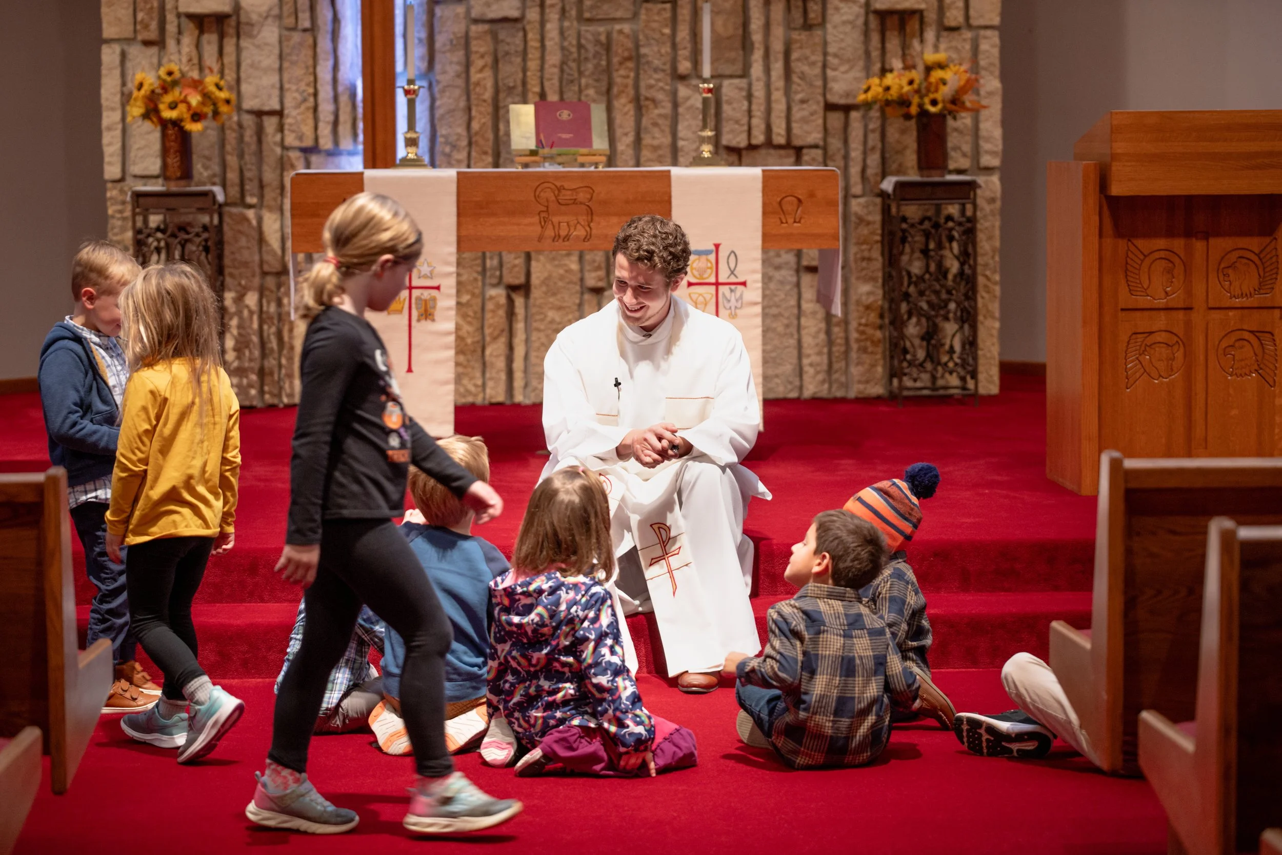 A priest in white robes kneels and smiles at a group of children sitting on red carpeted steps inside a church. Some children are standing and others are seated, with a few wearing rain gear. The church's altar with candles, a book, and floral arrangements in sunflowers is visible in the background.