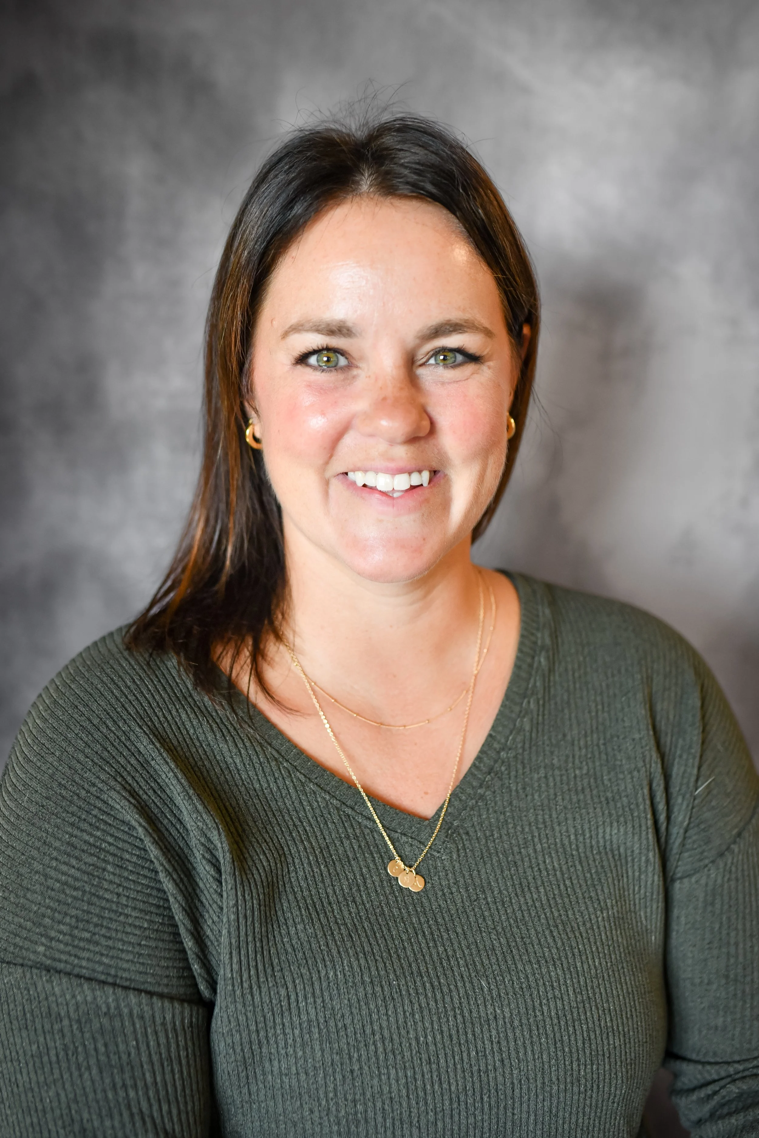 Portrait of a woman with dark brown hair, green eyes, wearing gold jewelry, smiling, against a plain gray background.