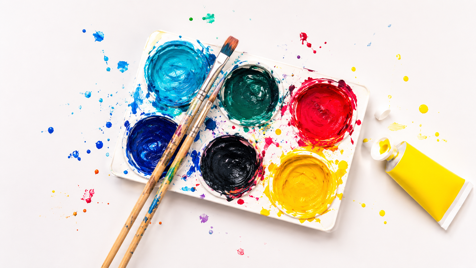 Paint palette with six colors of blue, green, red, dark blue, black, and yellow, along with two paintbrushes, some scattered paint drops, and a yellow tube of paint on a white surface.