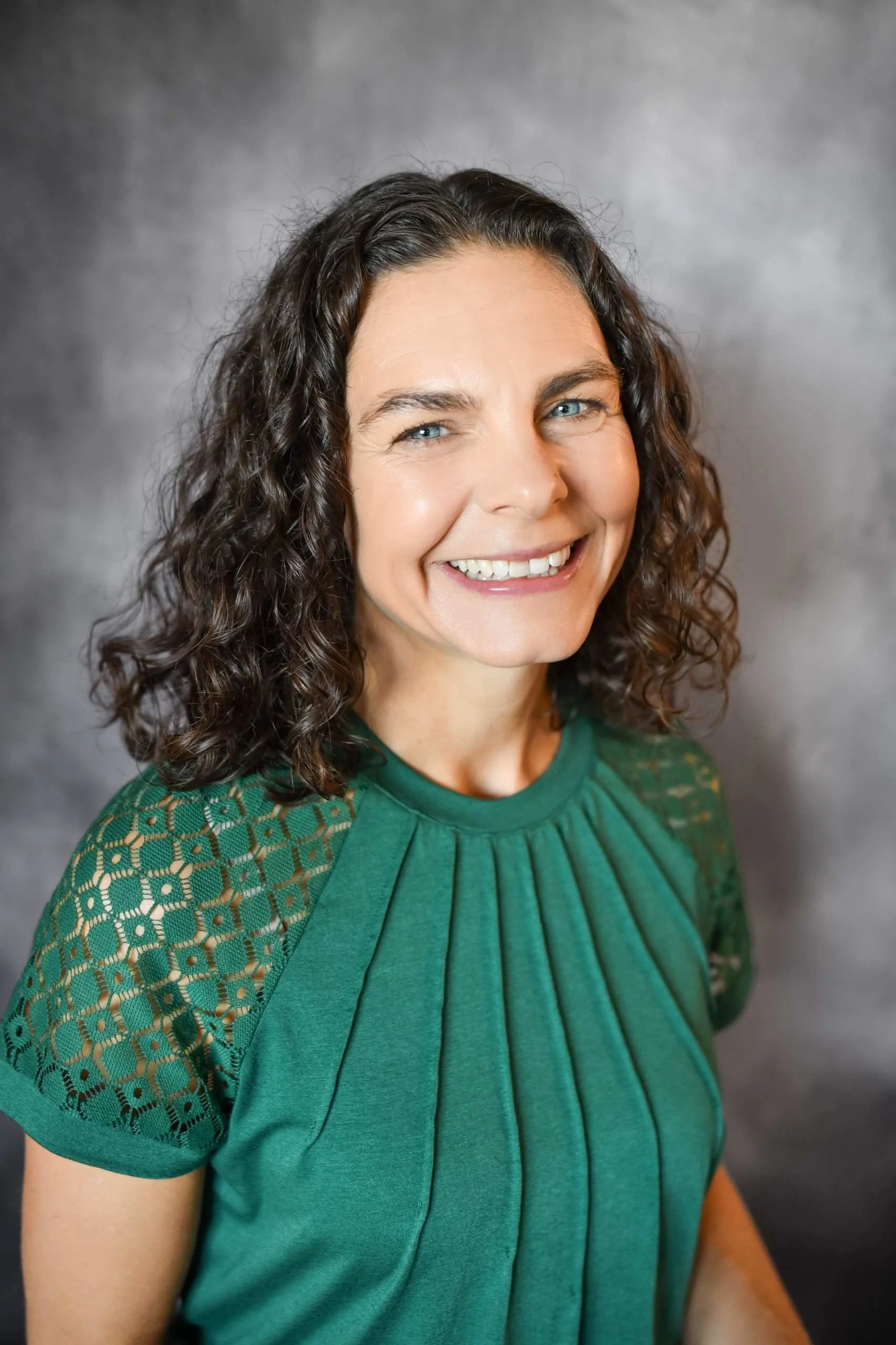Smiling woman with curly dark hair and blue eyes wearing a green top with lace details on the shoulders, against a gray background.
