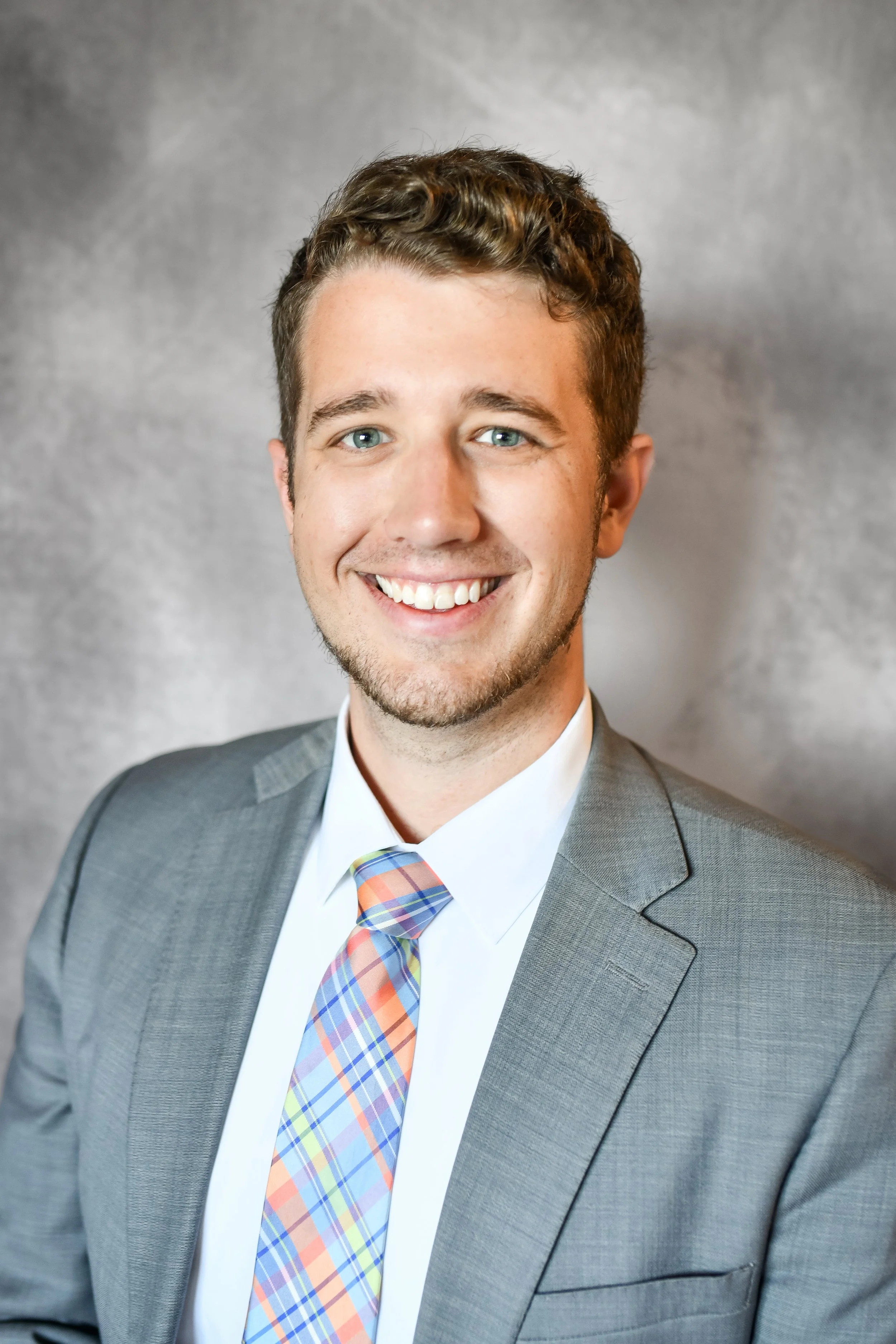 Young man with short brown hair, light skin, and blue eyes, smiling and wearing a gray suit with a white shirt and a colorful plaid tie, standing against a textured gray background.