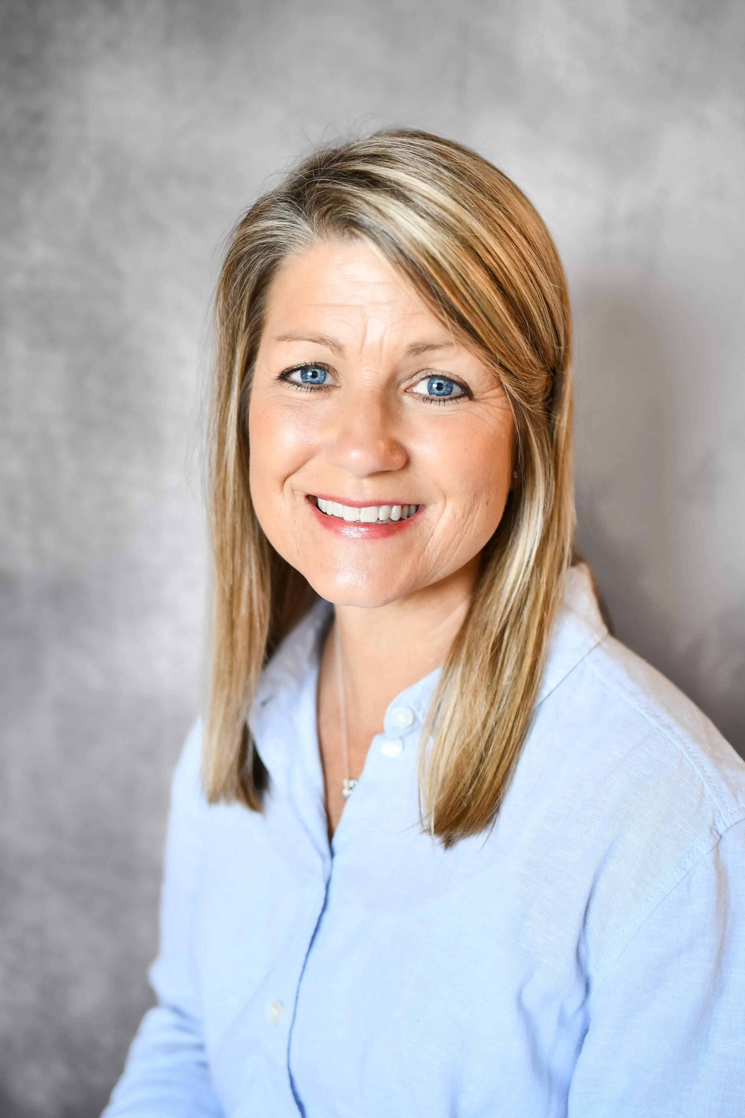 A woman with blonde straight hair and blue eyes smiling in a light blue collared shirt against a gray background.