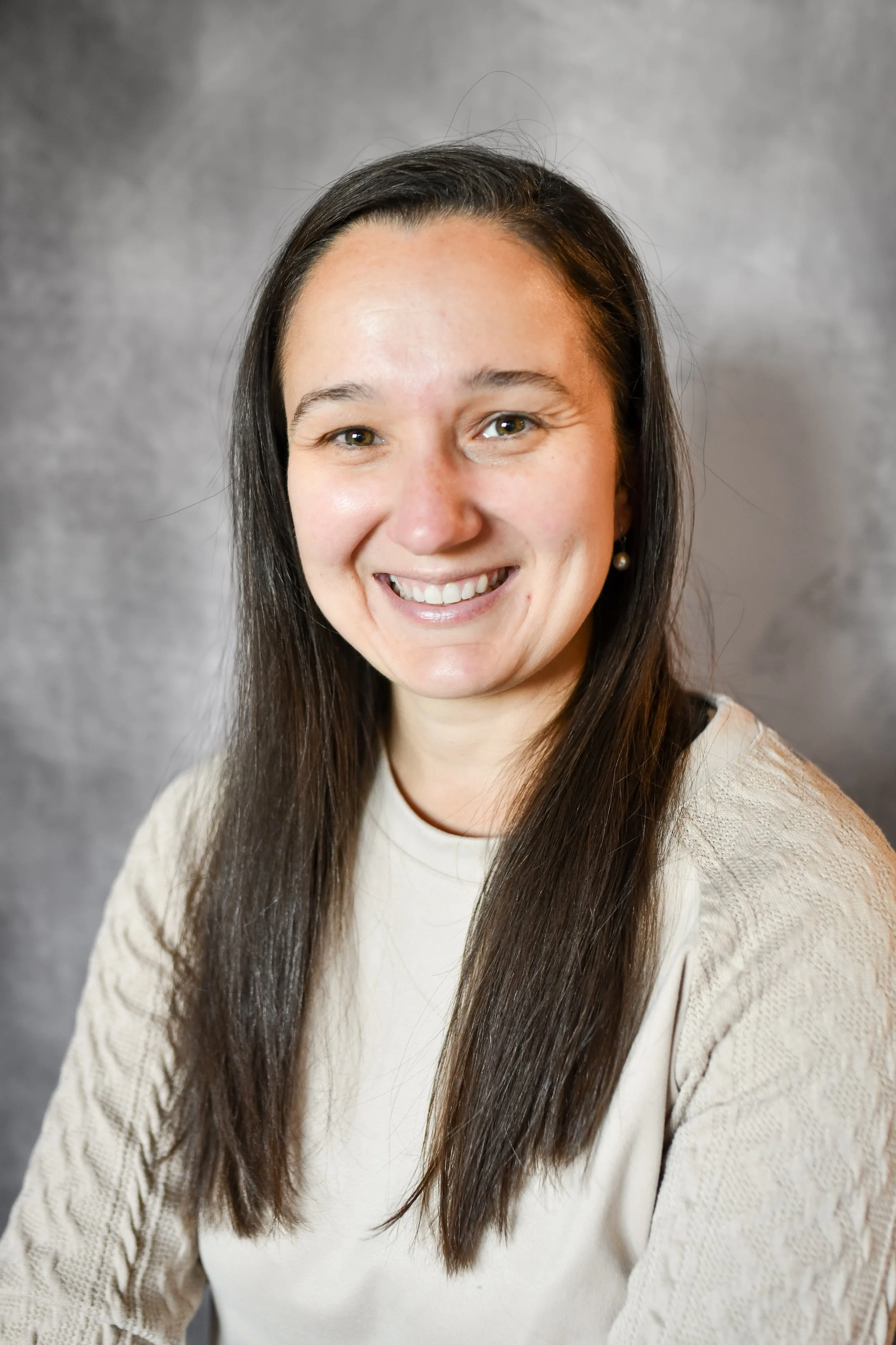 A woman with long dark hair, smiling, wearing a cream-colored sweater and earrings, against a gray background.
