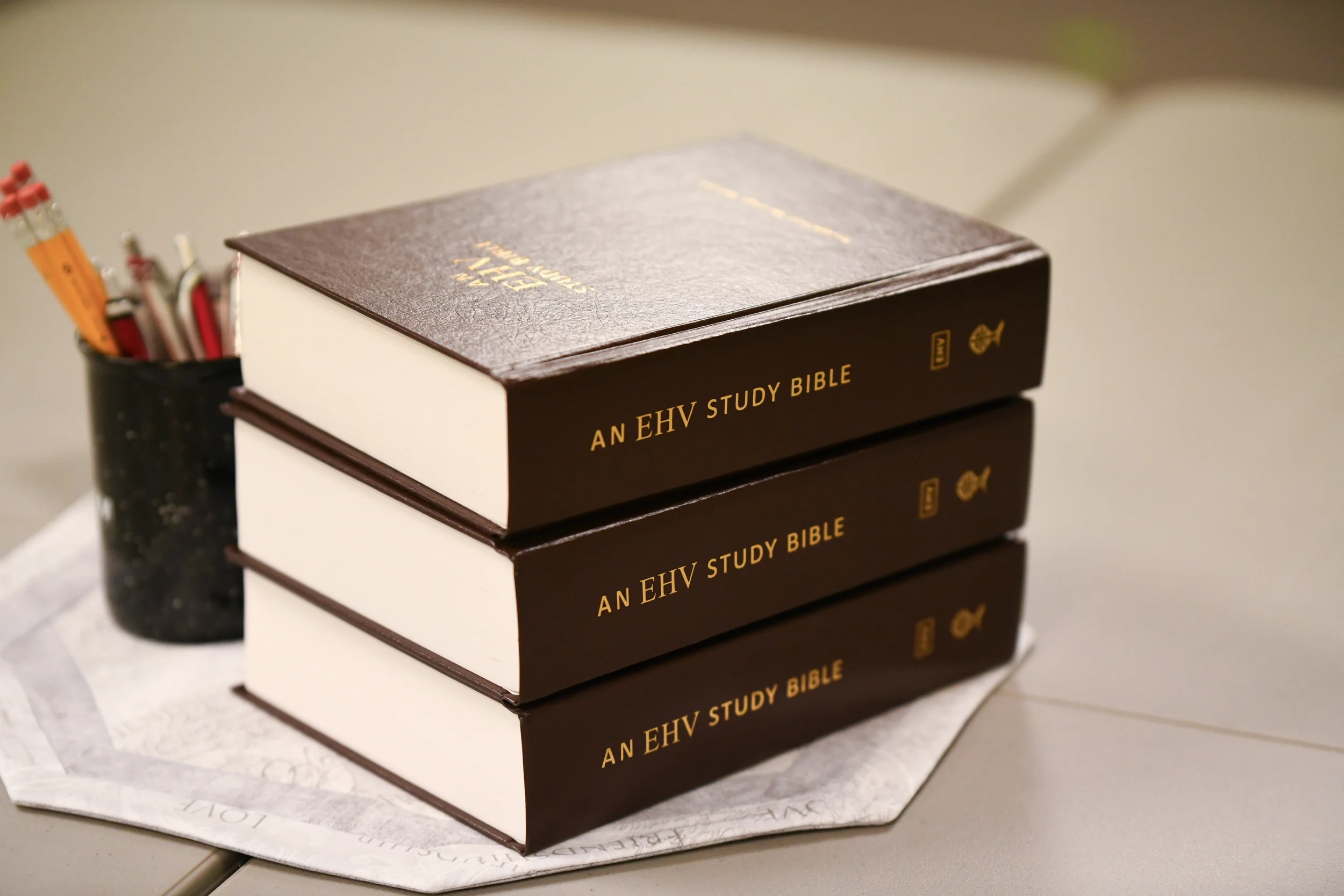 Stack of four black hardcover books titled 'An EHV Study Bible' with gold lettering, placed on a white paper wrapped around a marble surface, with a pencil holder containing pens and markers in the background.