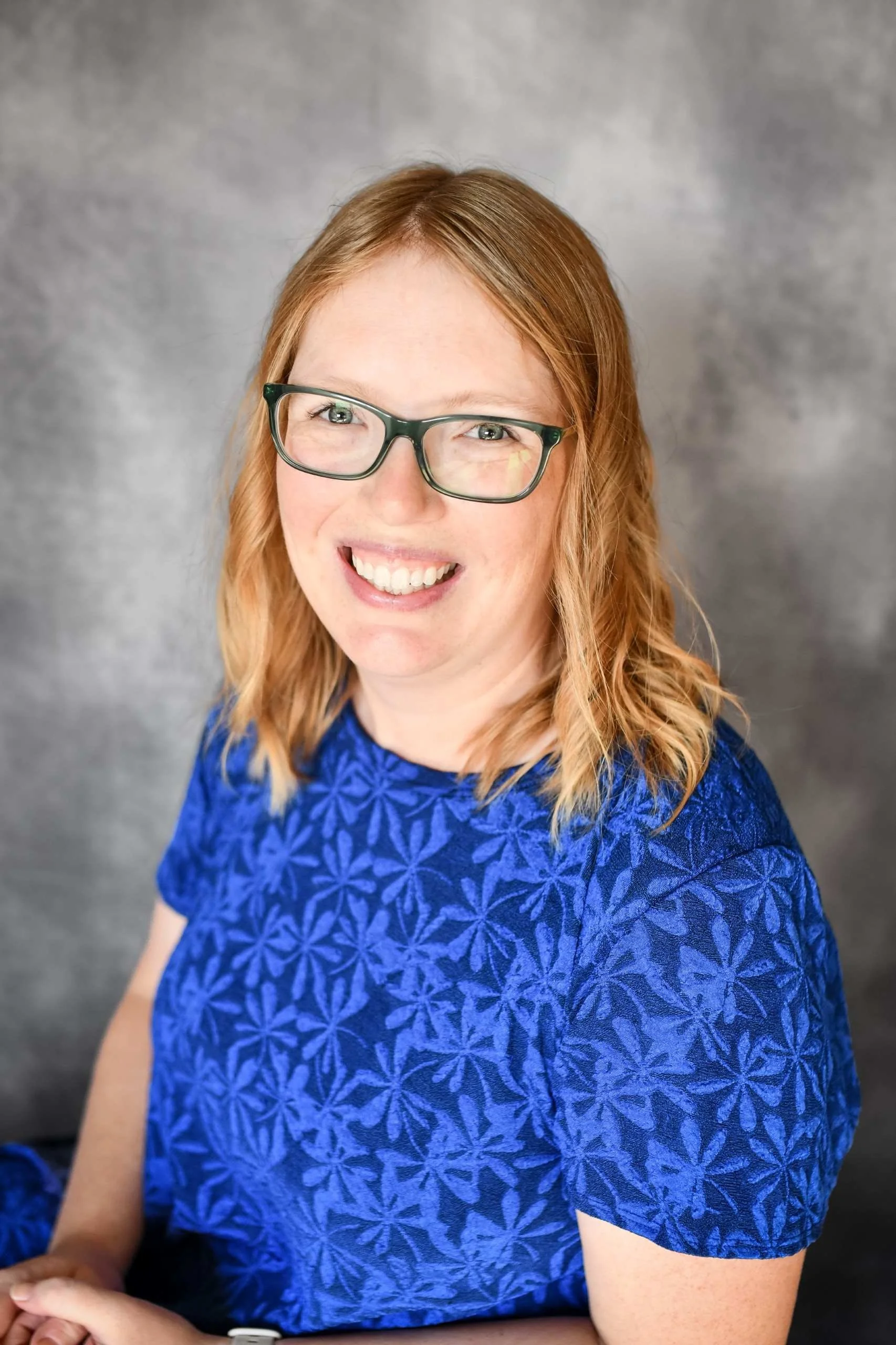 Close-up of a woman with red hair, glasses, smiling, wearing a blue patterned top, with a gray background.
