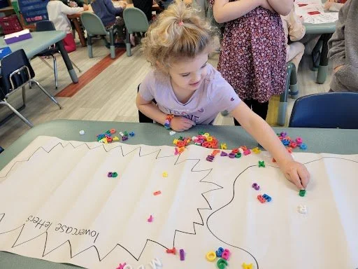 A young girl with blonde hair and a purple shirt decorating a large paper poster with colorful beads and craft supplies in a classroom or activity room.