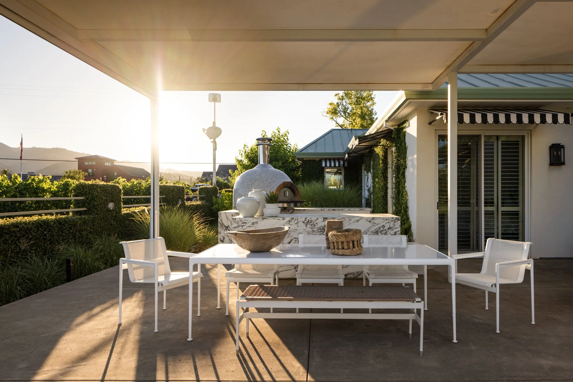 Outdoor patio with white table and chairs, a marble countertop with decorative bowls, and a pizza oven, illuminated by sunlight at sunset.