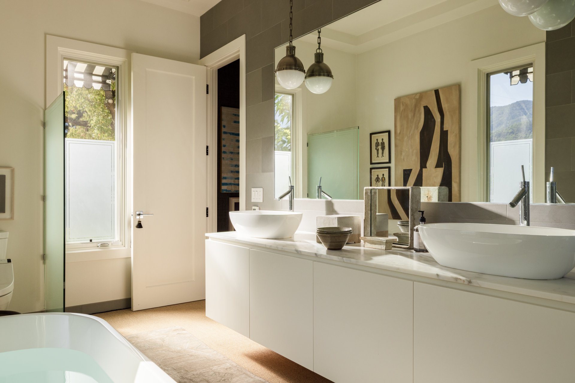 Modern bathroom with white fixtures, a marble countertop, wall-mounted faucets, and artwork on the wall. Two windows let in natural light, and a ceiling light fixture hangs above.
