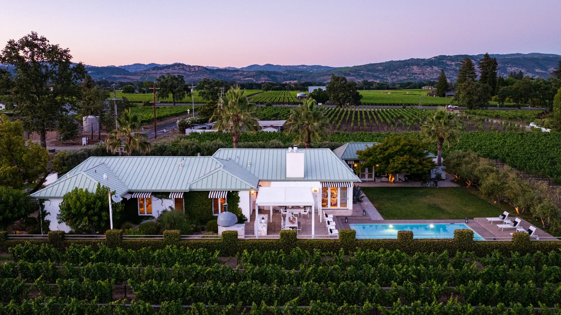 Aerial view of a house with a light-colored metal roof, a swimming pool, and outdoor seating area surrounded by landscaped gardens and lush greenery. Vineyard rows and rolling hills are visible in the background at sunset.