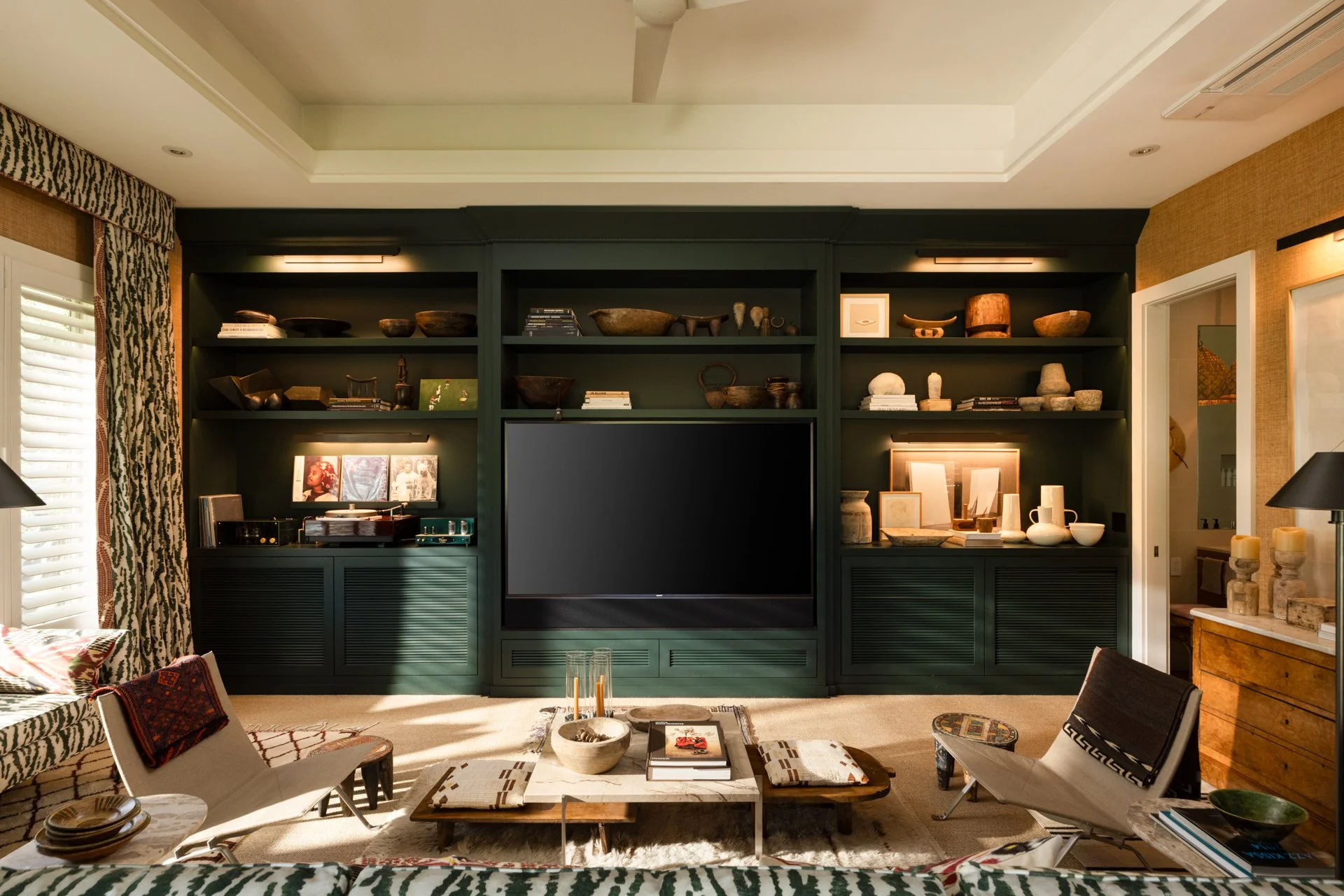 Living room with dark green built-in bookshelf containing decorative bowls, books, and framed artwork, with a large flat-screen TV in the center, and eclectic furniture including a marble coffee table, patterned cushions, and modern chairs.