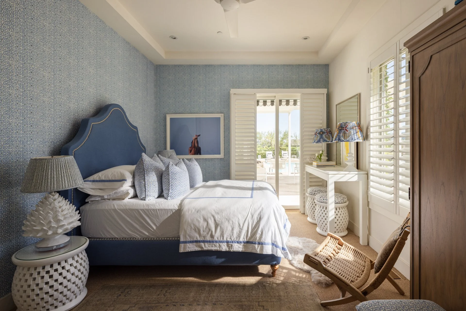 A bedroom with a blue upholstered headboard, a white bed with decorative pillows, a white side table with a textured table lamp, a wall painting, a white vanity table with a stack of books and two patterned lamps, two white woven stools, a wooden arm