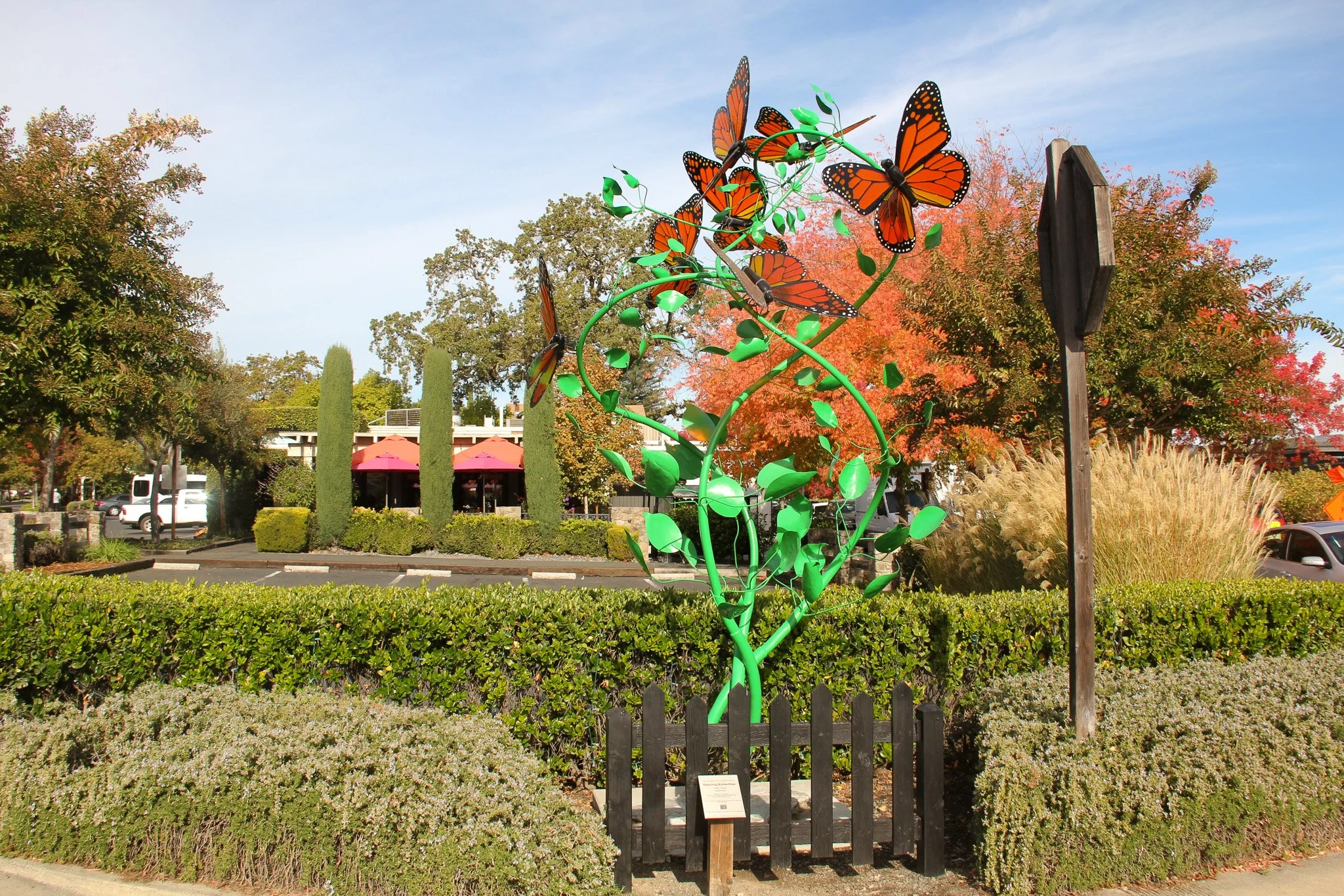 A colorful outdoor sculpture of orange and black butterflies on green branches in a park with trees and a parking lot in the background.