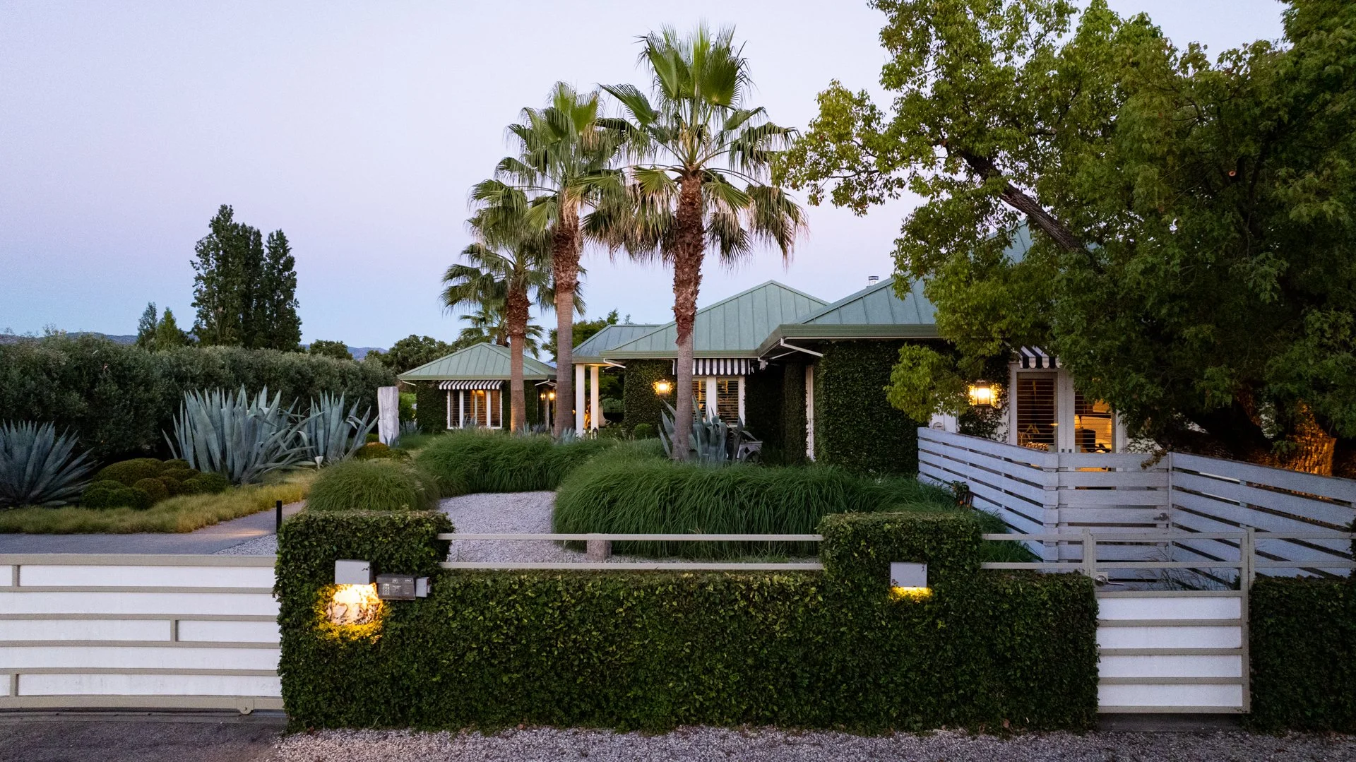 A suburban house with a garden, tall palm trees, and a white fence in the foreground during evening, with the house lights on.