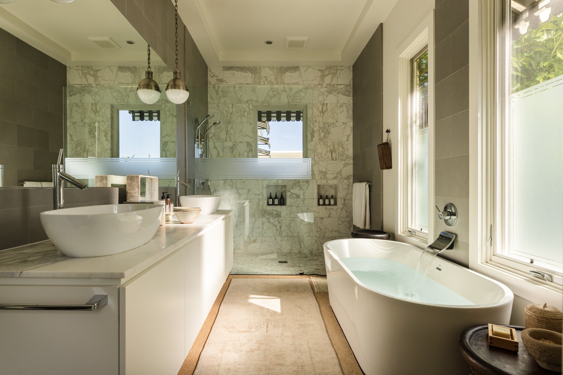 Bathroom with a white bathtub, marble walls, a large mirror, a double sink, and large windows letting in natural light.