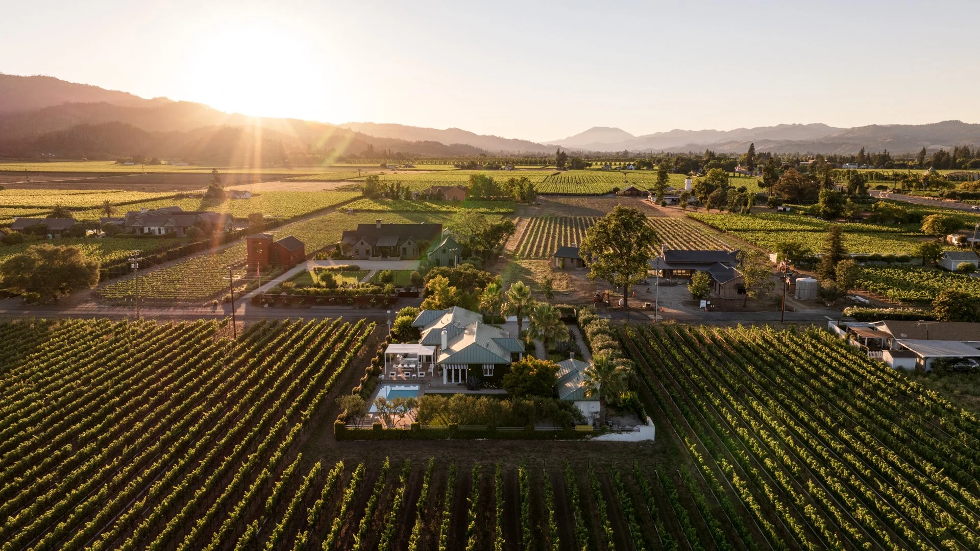 Sunset over an expansive vineyard landscape with scattered houses, trees, and mountains in the background.