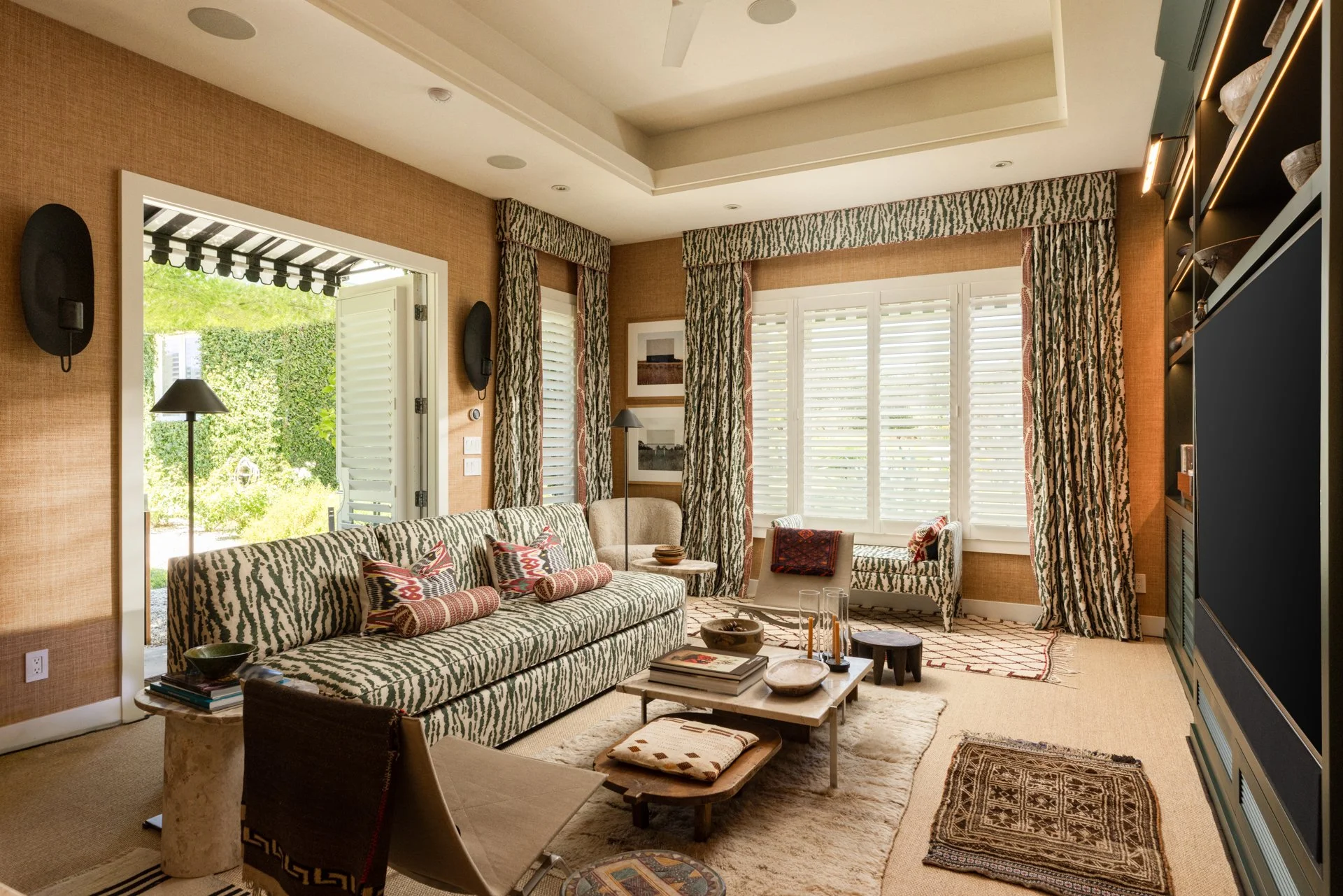 Living room with patterned sofa, armchair, coffee table, and large windows with zebra-striped curtains, open to a garden patio.