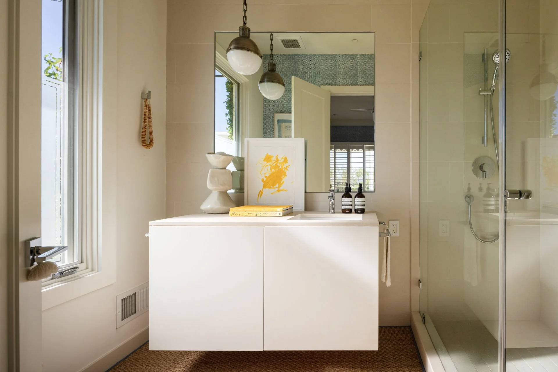 A modern bathroom with a white vanity, decorative items, and a glass shower. There are two pendant lights hanging from the ceiling, a window with white blinds, and a large mirror above the vanity. The walls are a light color, and there's a small rug 