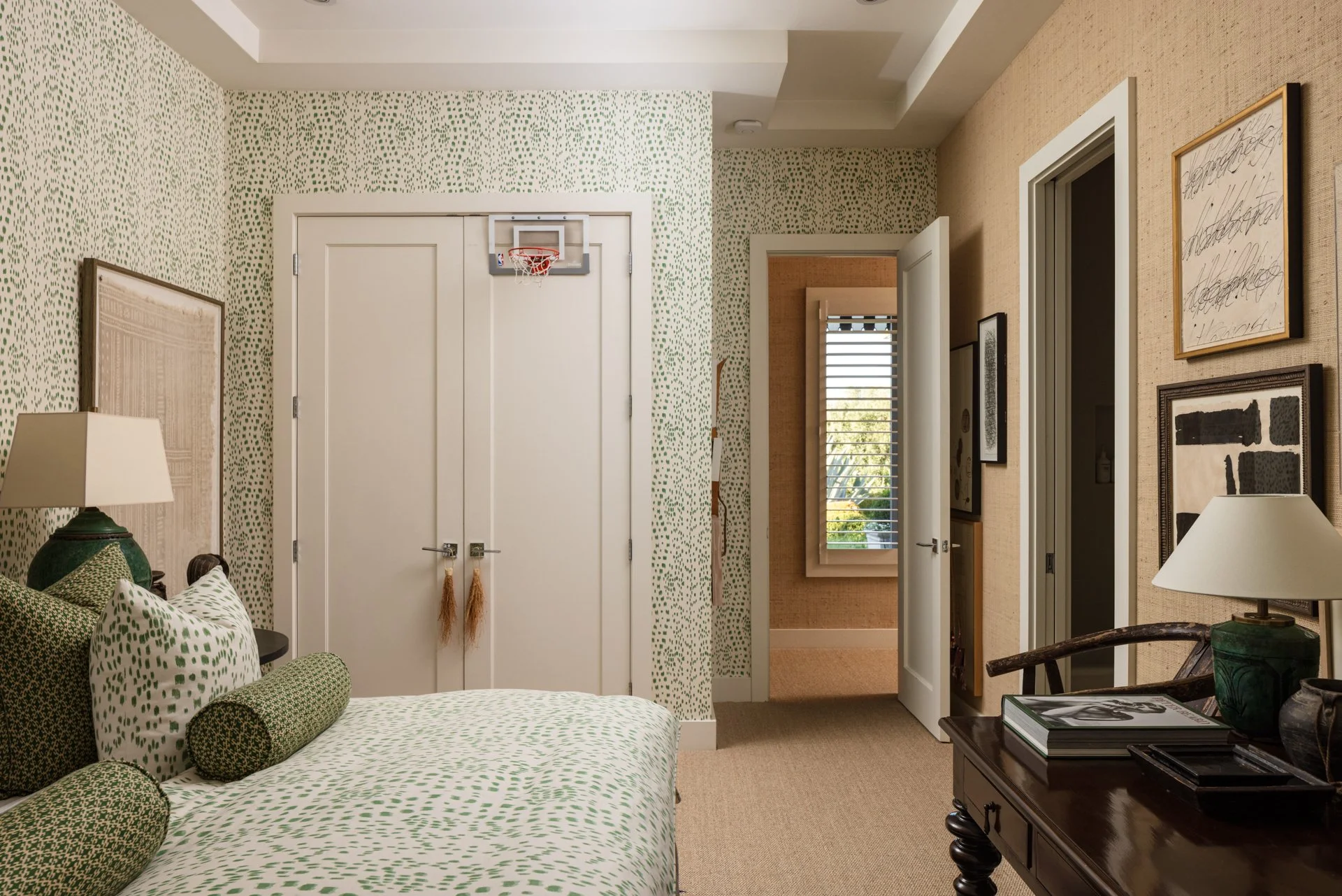 An interior room with patterned wallpaper, a bed with green and white bedding, portraits on the wall, a bedside table with a lamp, and a window with blinds.