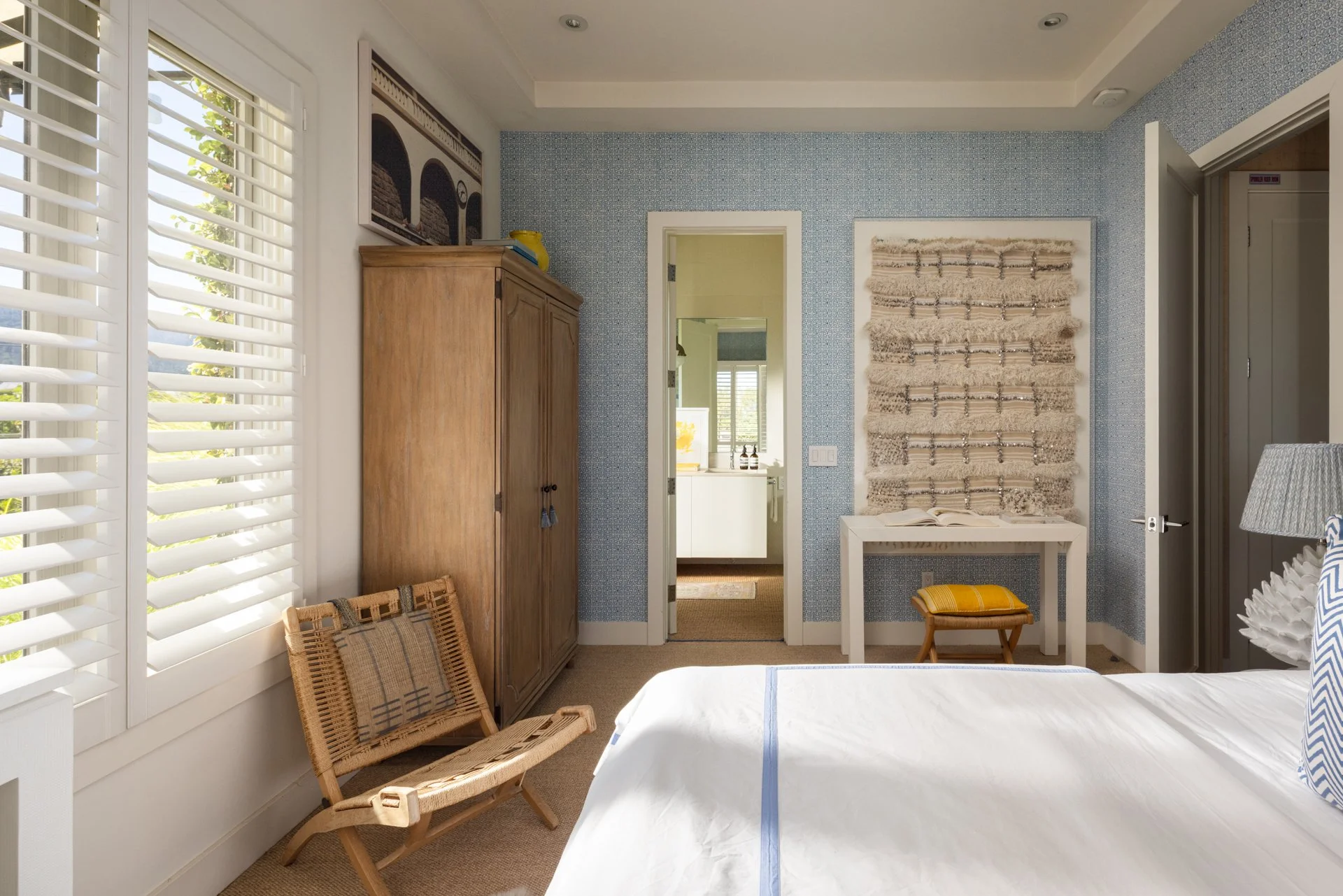 A cozy bedroom with large white plantation shutters on the window, a woven wooden chair with a plaid cushion, a tall wooden wardrobe, and a doorway leading to a bright bathroom. The wall is painted with blue patterned wallpaper and has a textured wal