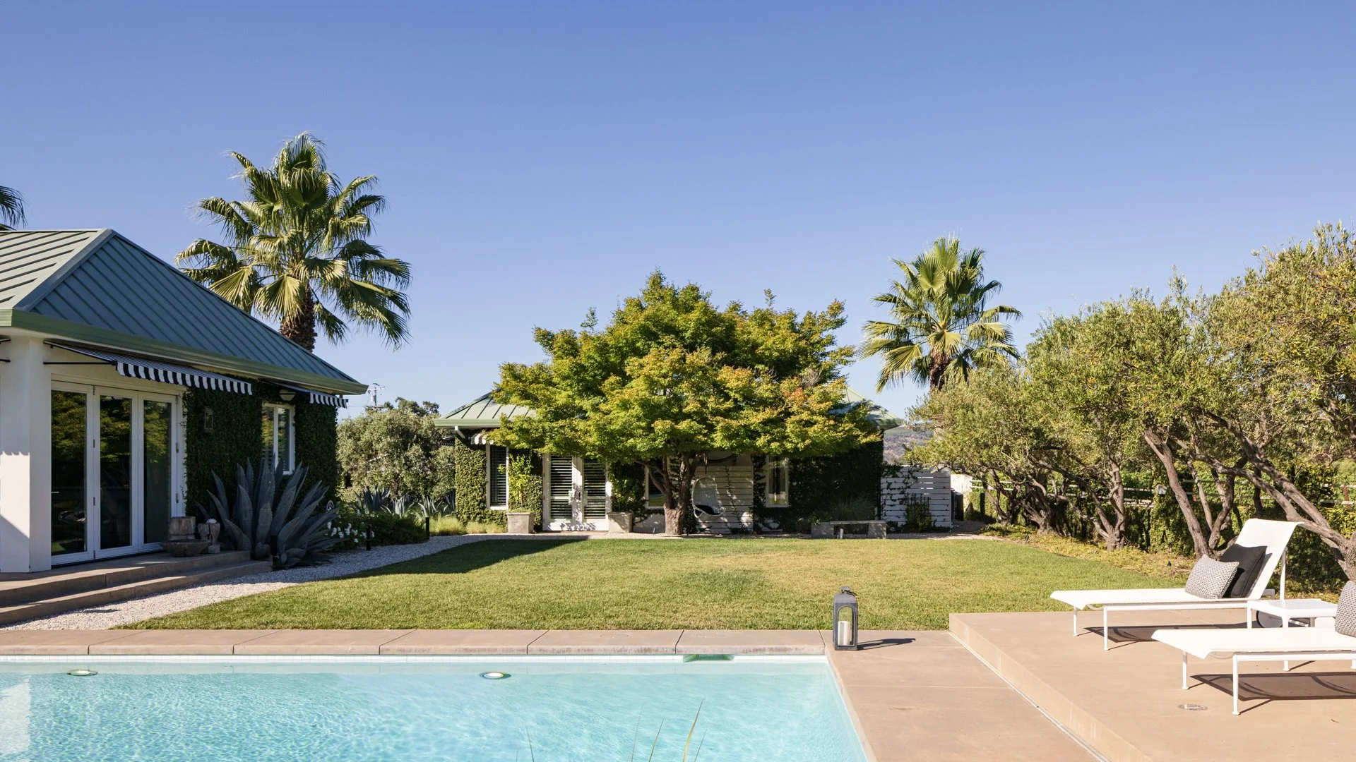 Backyard with a swimming pool, patio chairs, trees, and a house with a green metal roof and sliding glass doors.