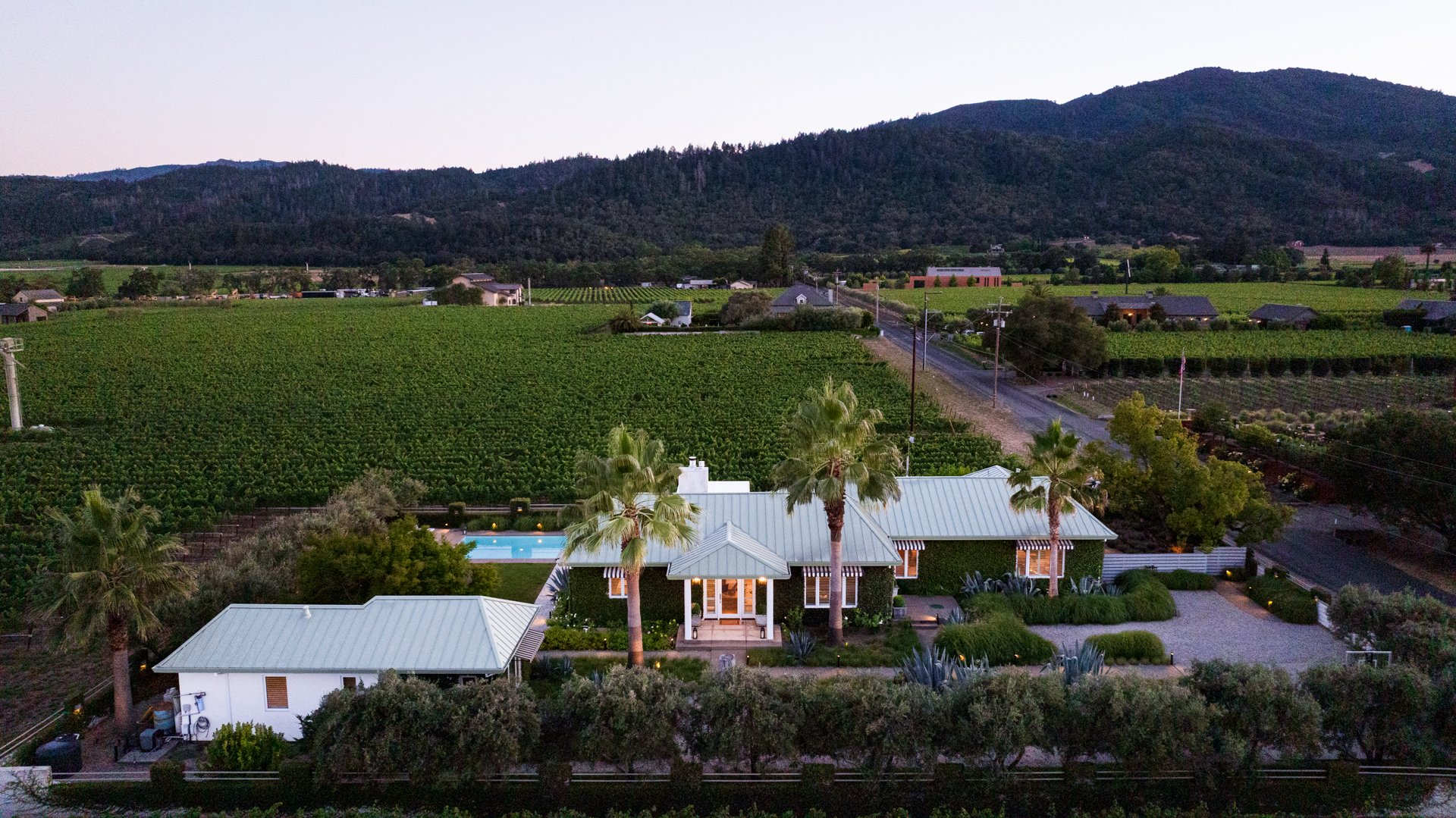 Aerial view of a rural landscape at dusk featuring a white house with a metal roof, palm trees, a swimming pool, green fields, and rolling mountains in the background.