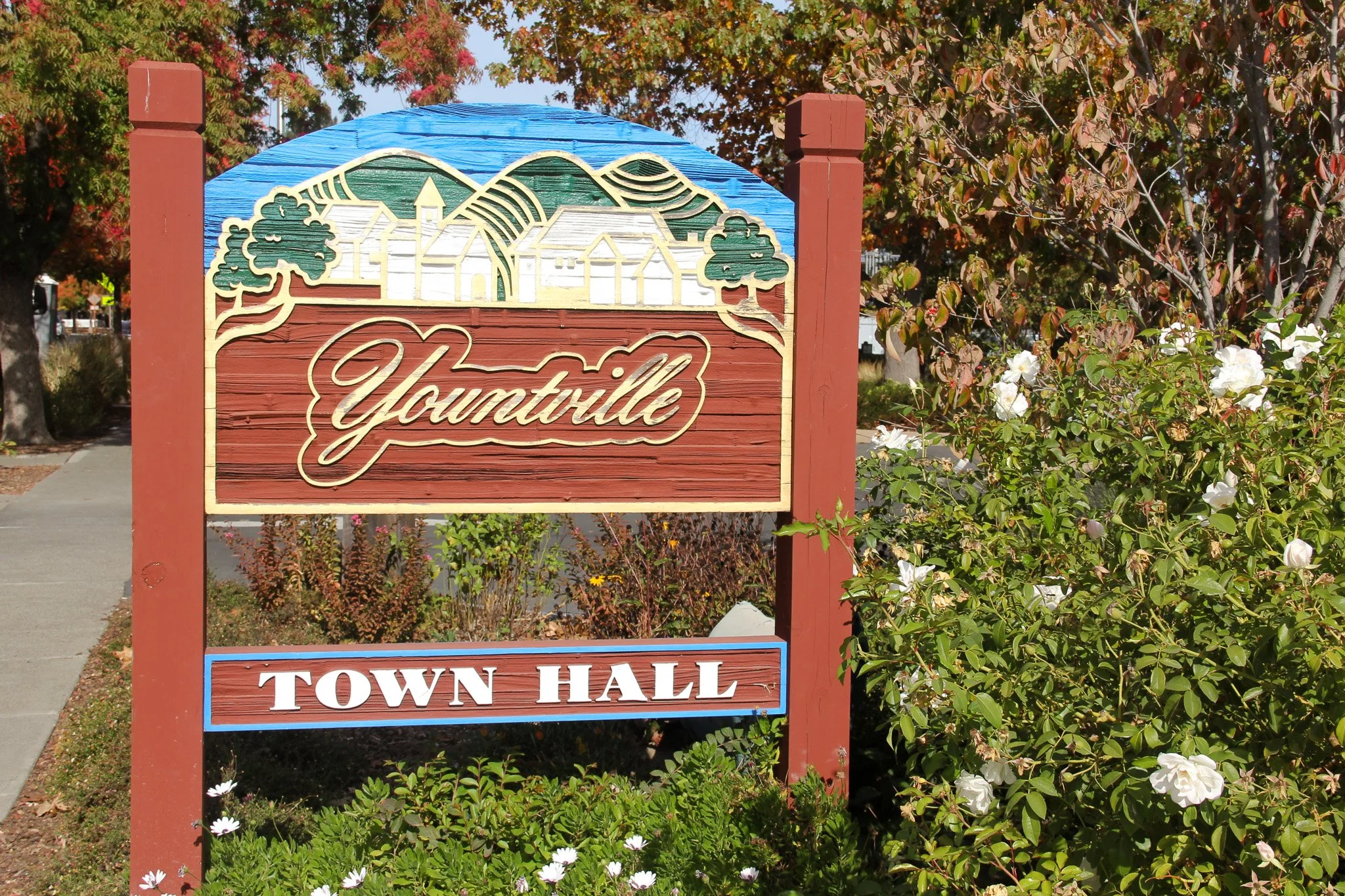 A colorful sign with a mountain, trees, and houses illustration reads 'Yountville' and 'Town Hall' with surrounding bushes and flowering plants, trees in the background.