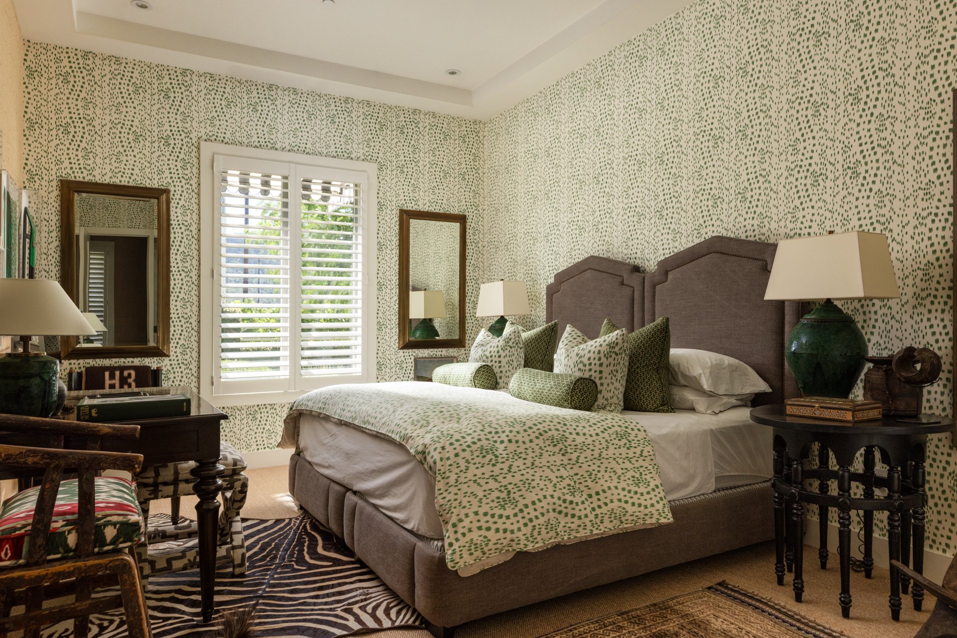 A bedroom with green and white dotted patterned walls, a bed with matching bedding and pillows, two nightstands with lamps, and a window with plantation shutters.
