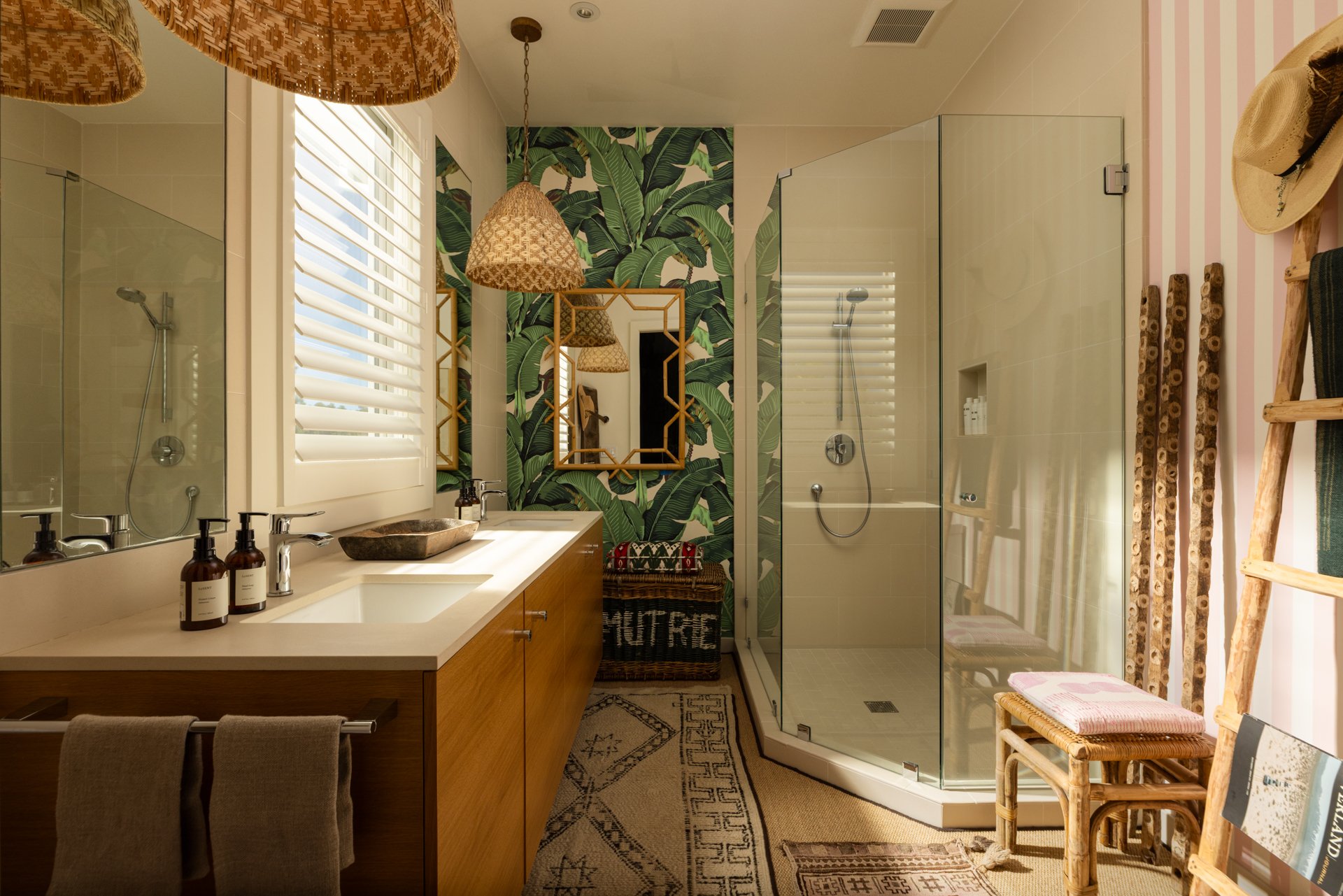 A bathroom with a large mirror above a white countertop featuring a stone sink, surrounded by bottles for soap or lotion. The background includes a shower with a glass door, tropical leaf wallpaper, a woven basket, and a small bench with towels. Ther